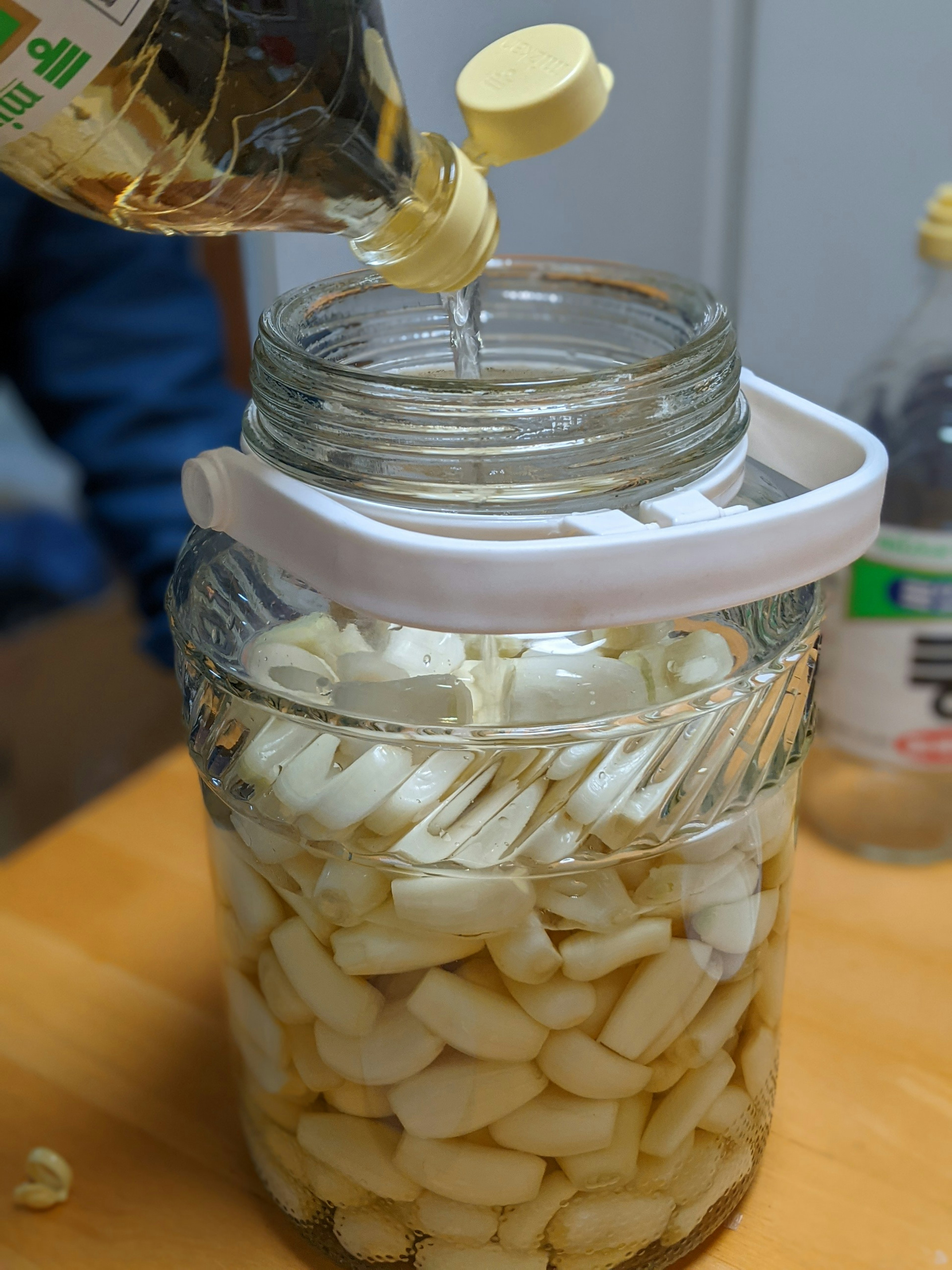 A large glass jar filled with peeled garlic cloves as oil is being poured in