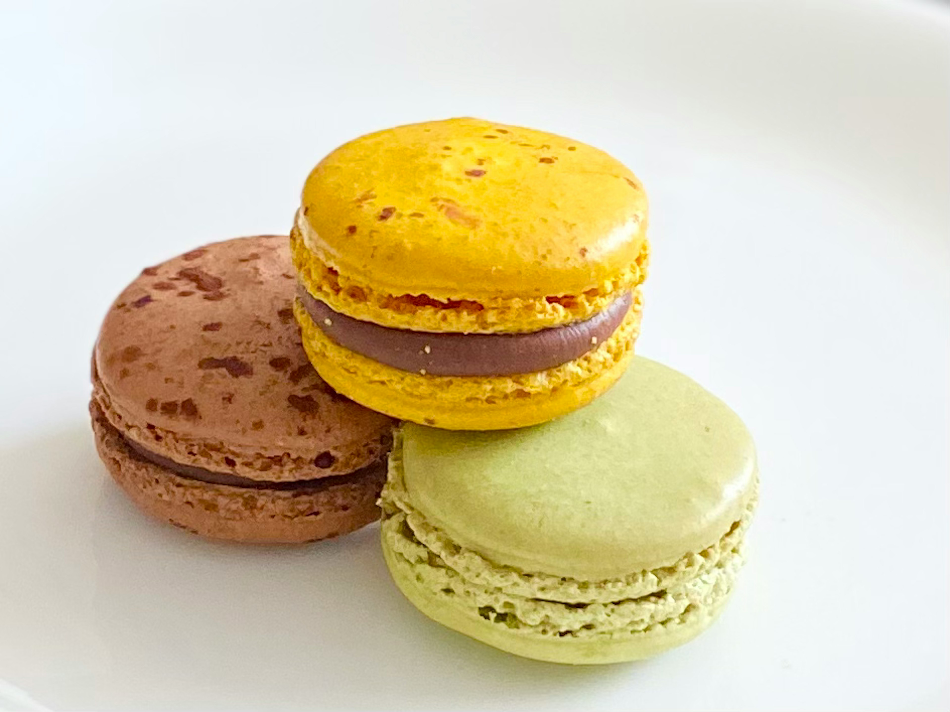 Tiga macarons di atas piring putih berwarna coklat, kuning, dan hijau dengan isian yang terlihat