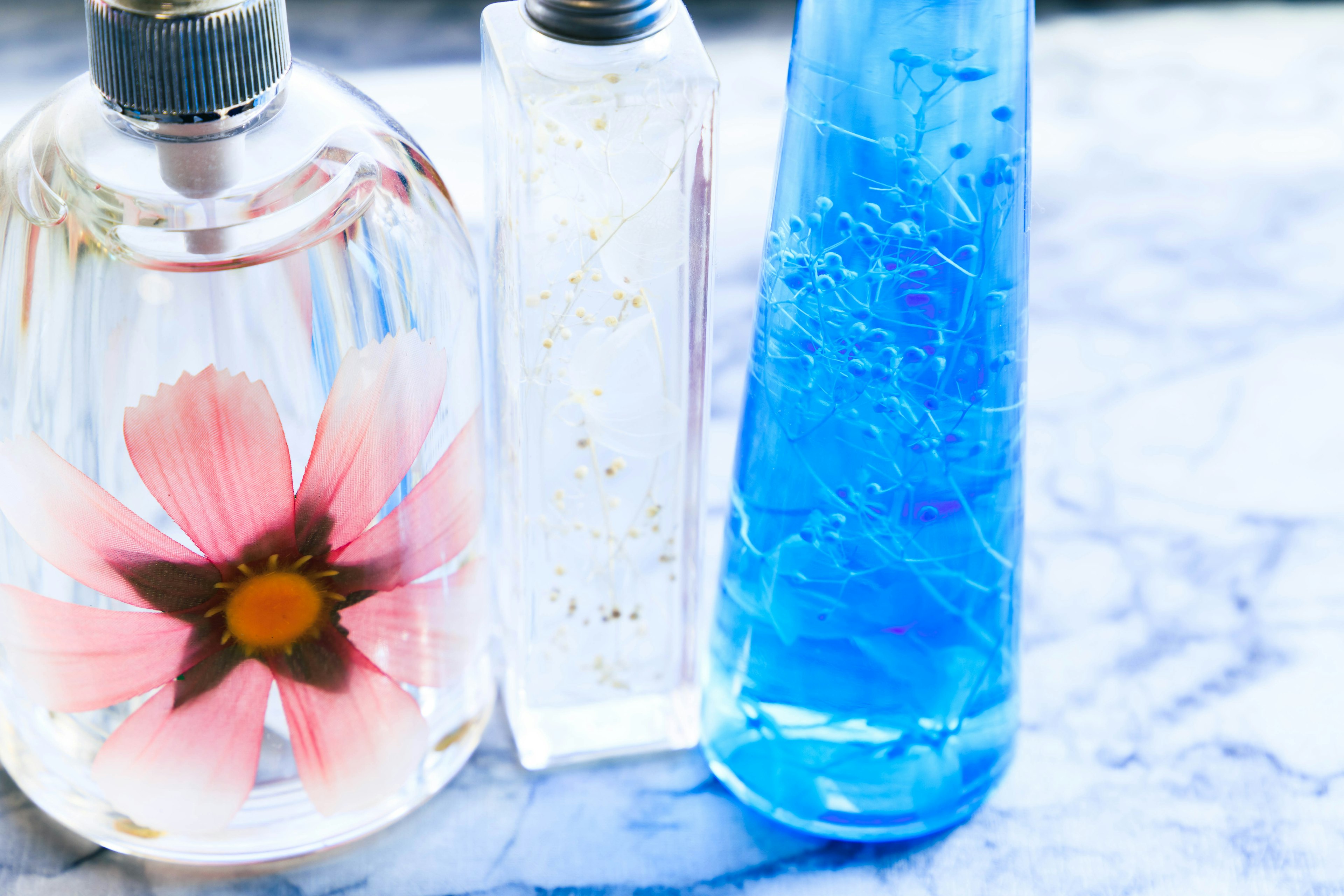 Tres botellas decorativas con líquidos florales y azules sobre una superficie de mármol