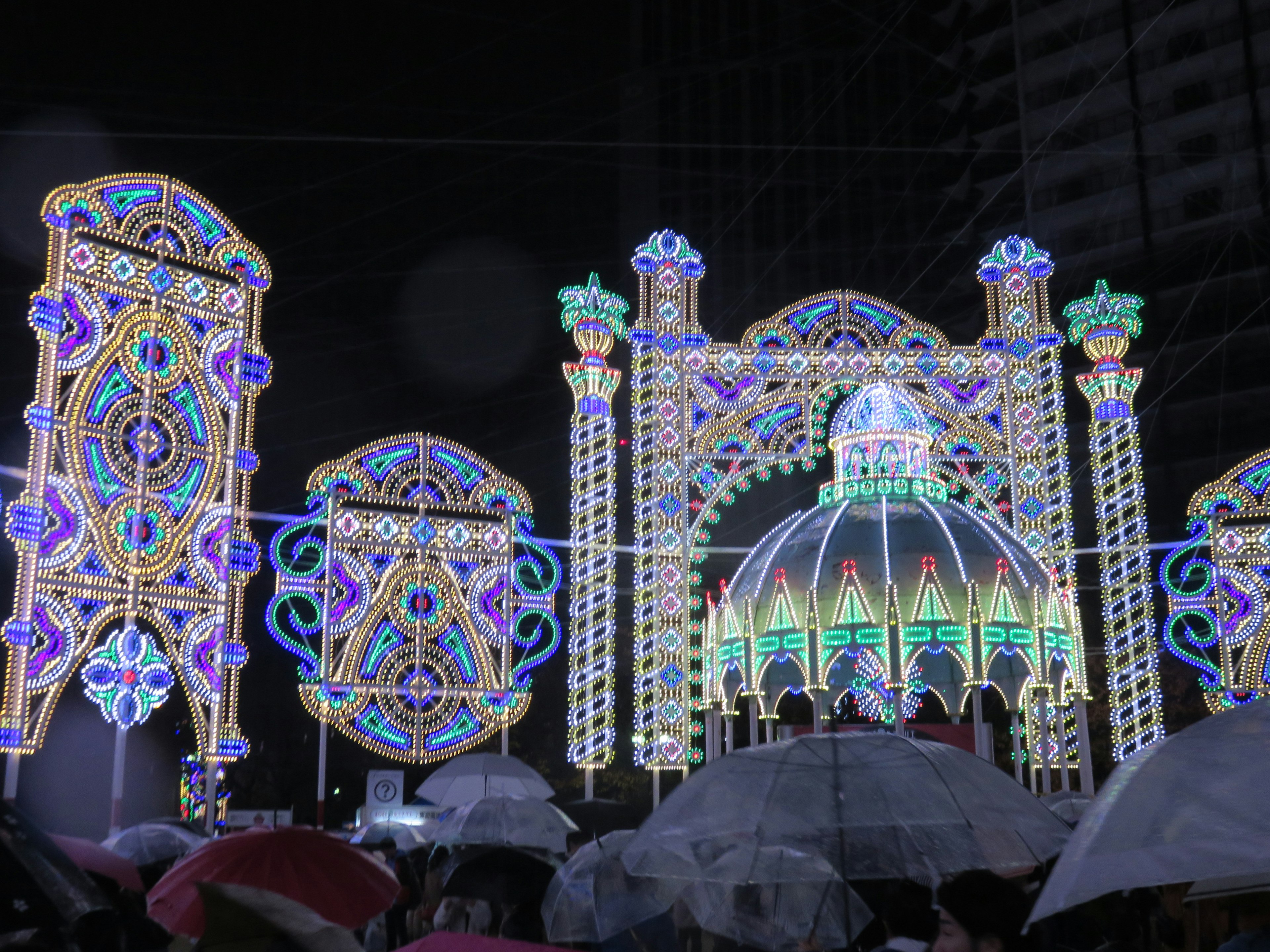 Colorful illuminated arch and dome in a night scene with people holding umbrellas