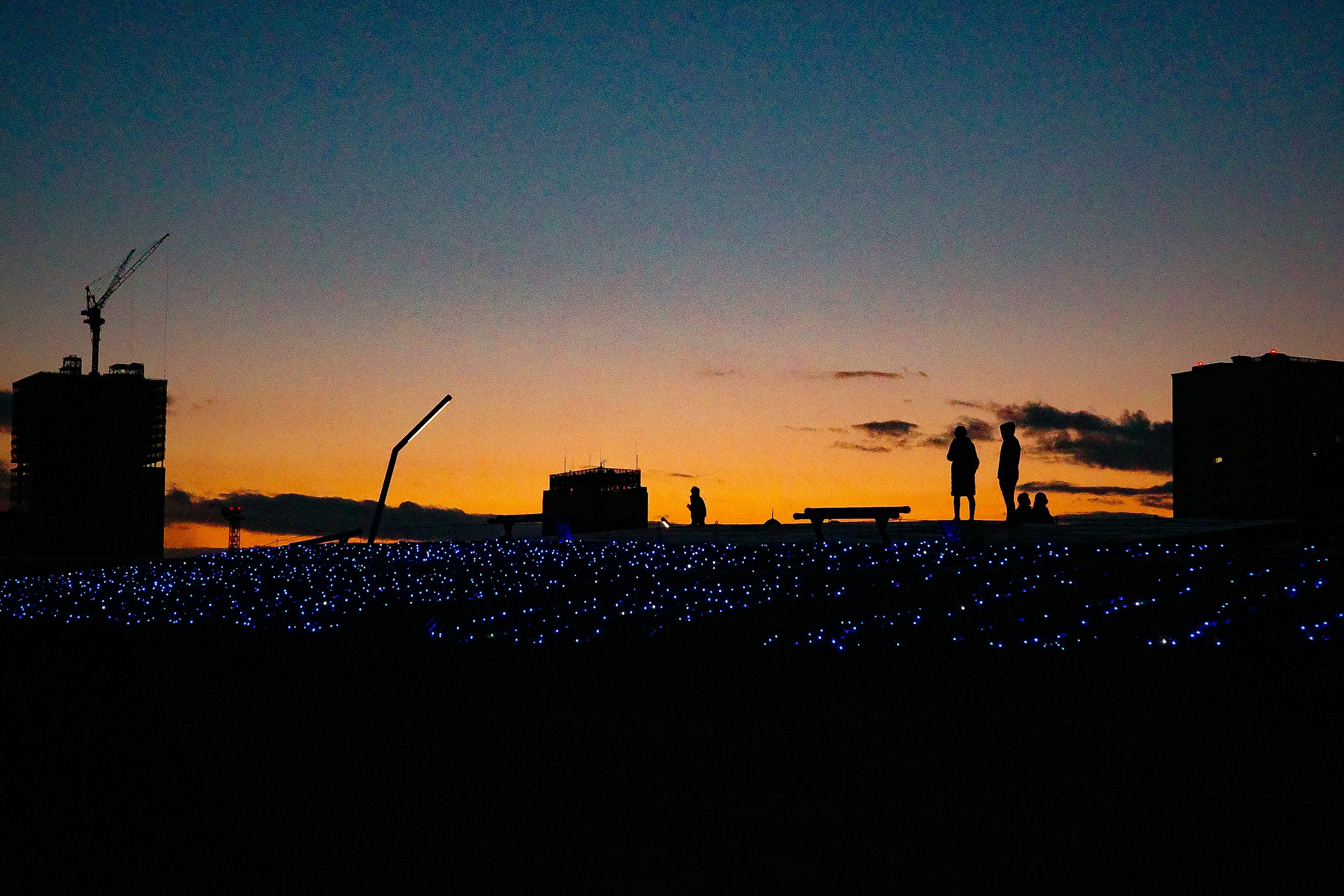 夕焼けの中、青い光の海が広がり、シルエットの人々が立っている風景