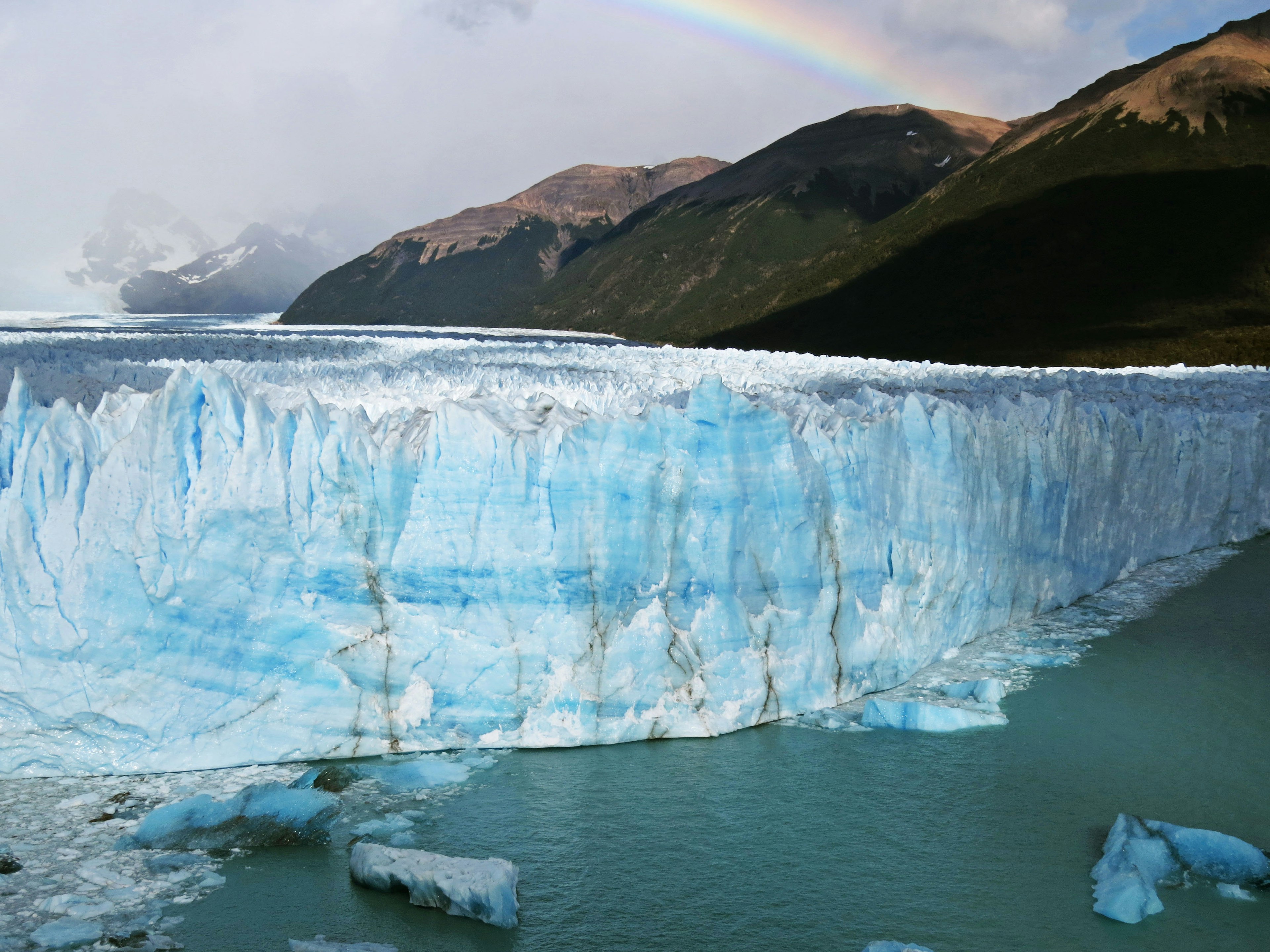 Pared de hielo azul de un glaciar con montañas y un arcoíris