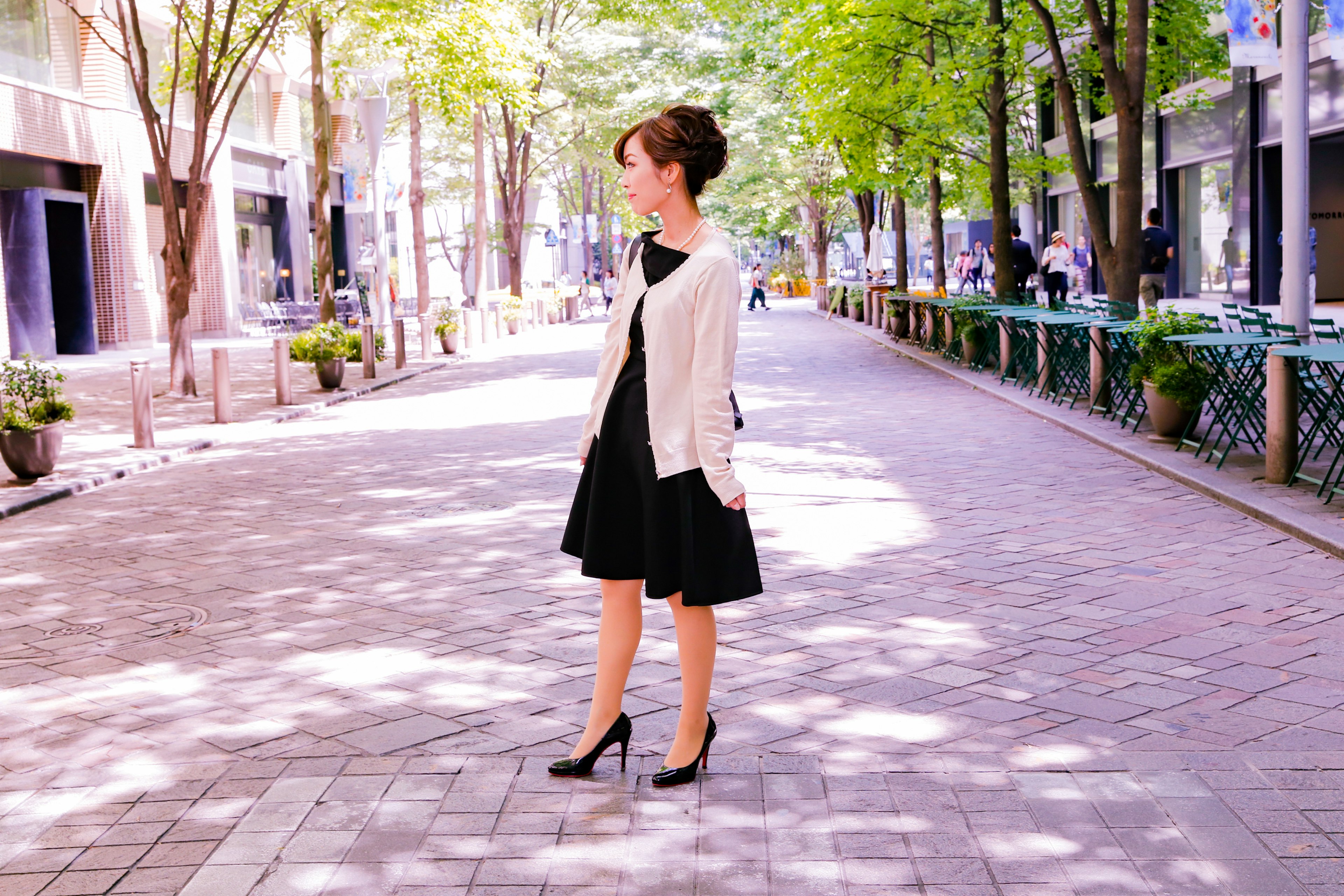 A woman in a black dress stands on a sunny street