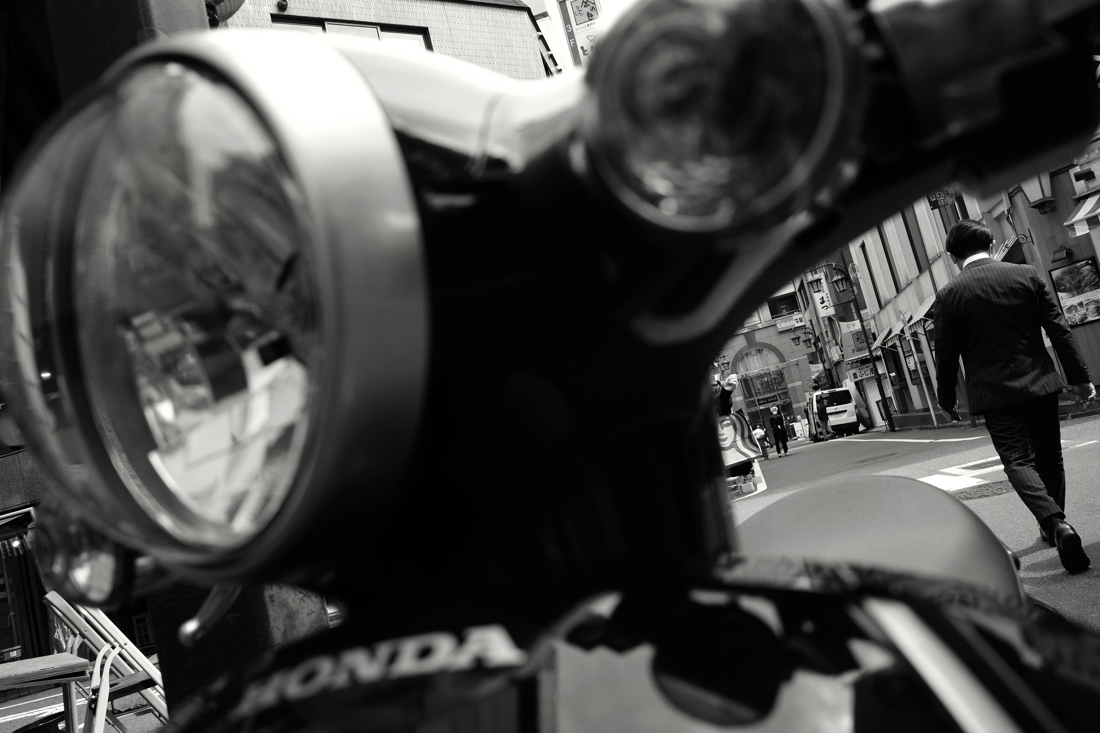 Close-up of a motorcycle headlight and handlebars with a person walking in the background