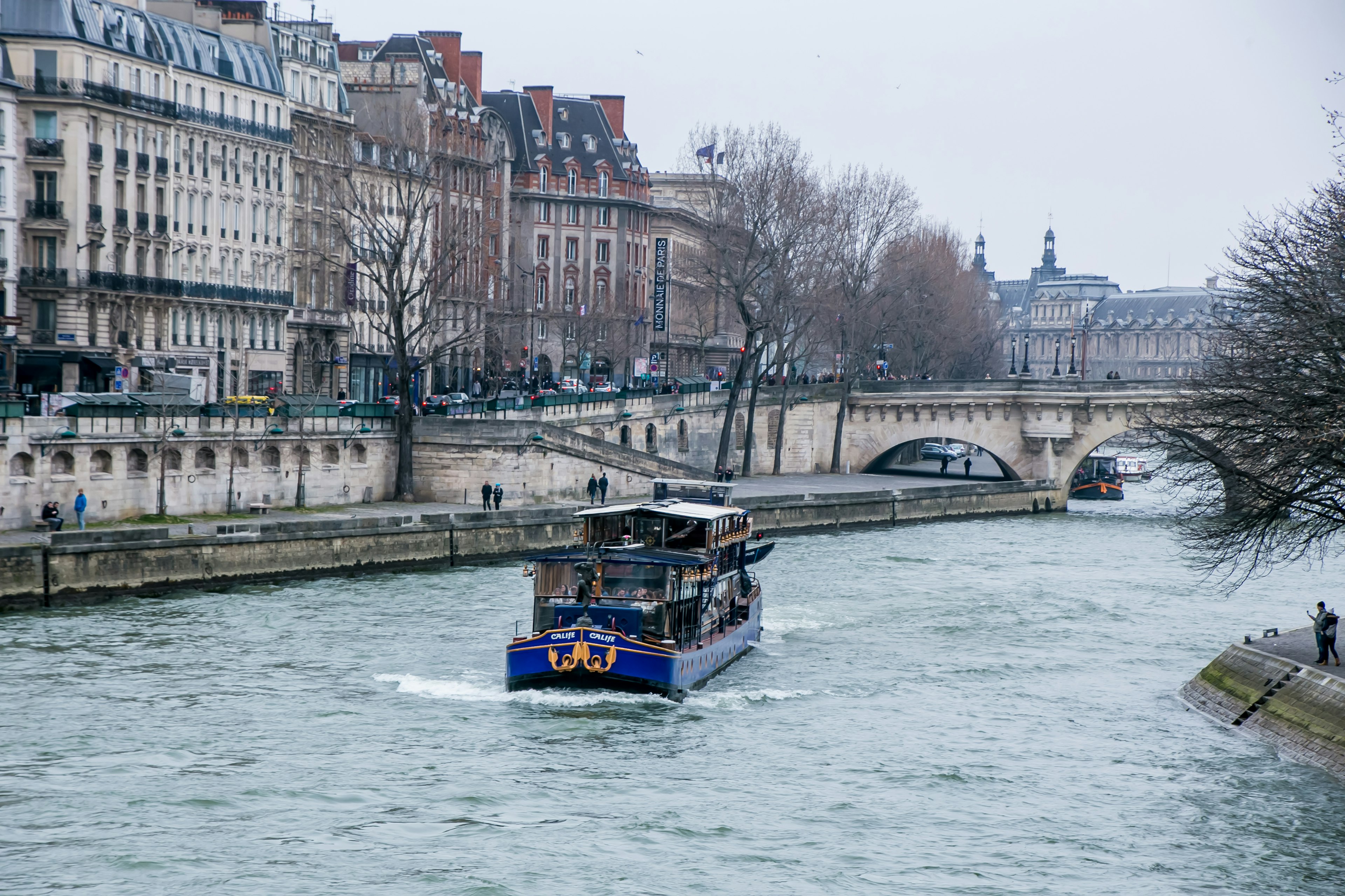 Thuyền màu xanh di chuyển trên sông Seine với các tòa nhà Paris