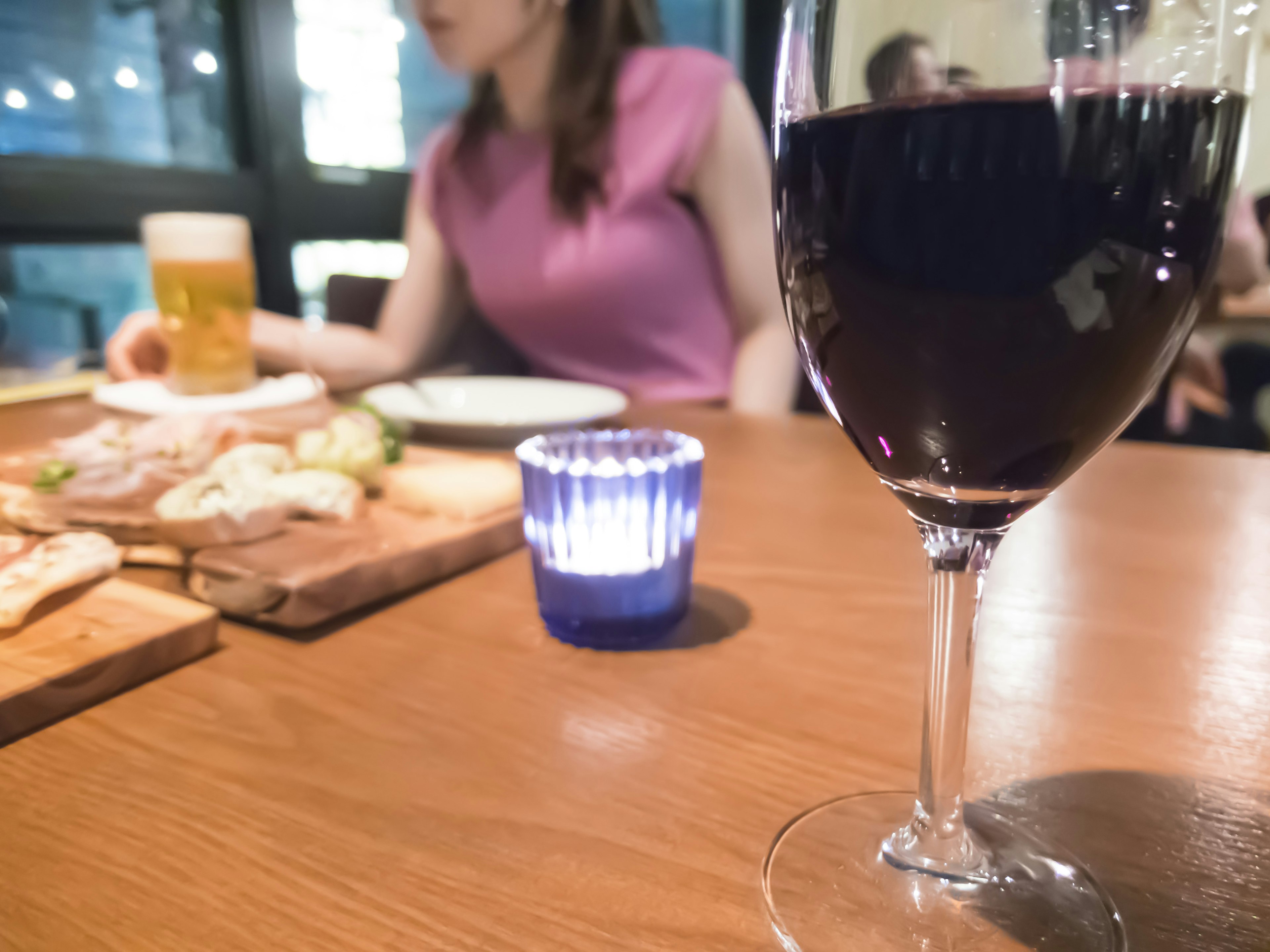 A glass of red wine on a table with food and a woman seated in the background