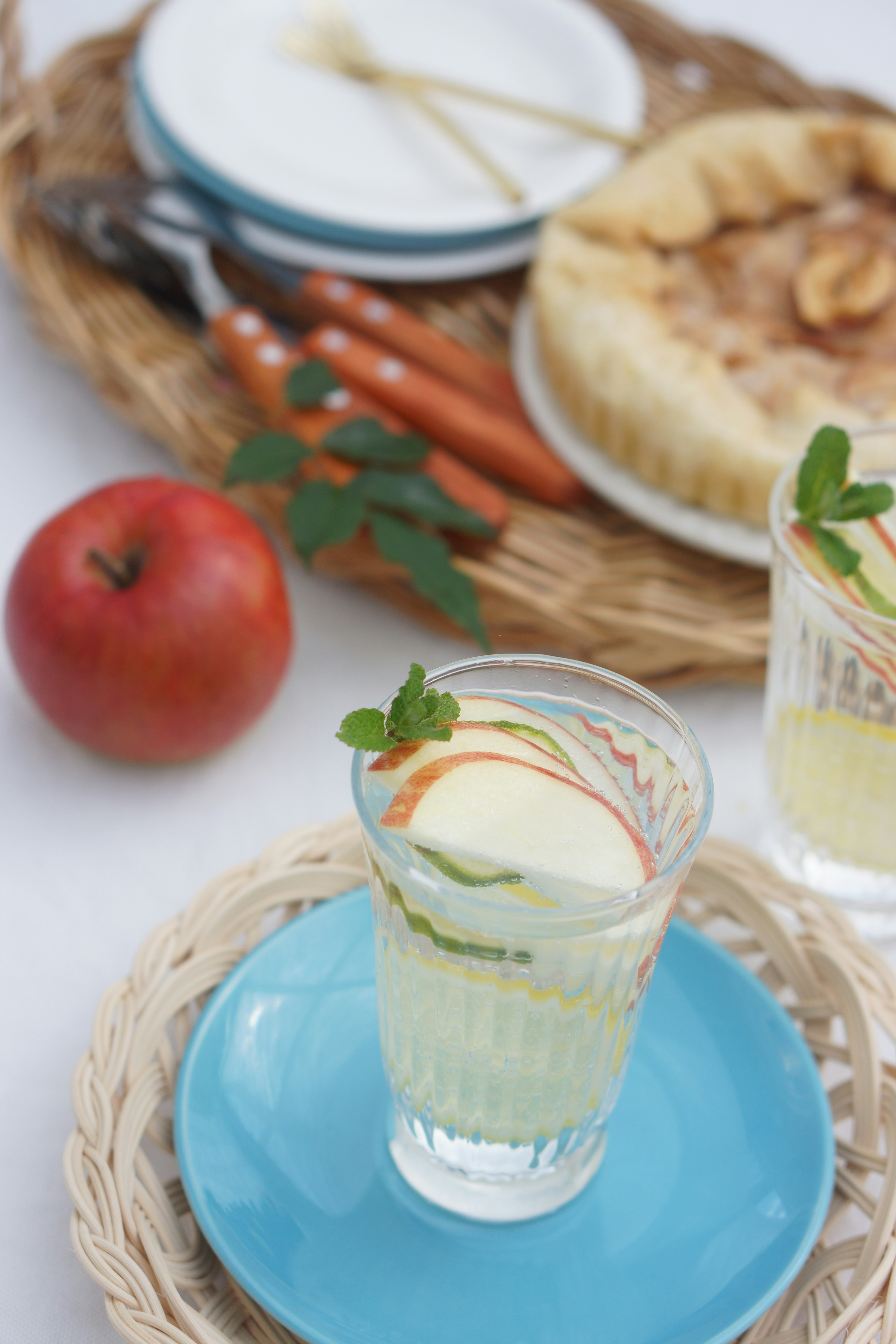 Une boisson avec des tranches de pomme sur une assiette bleue à côté d'une pomme et d'une tarte sur un plateau tissé