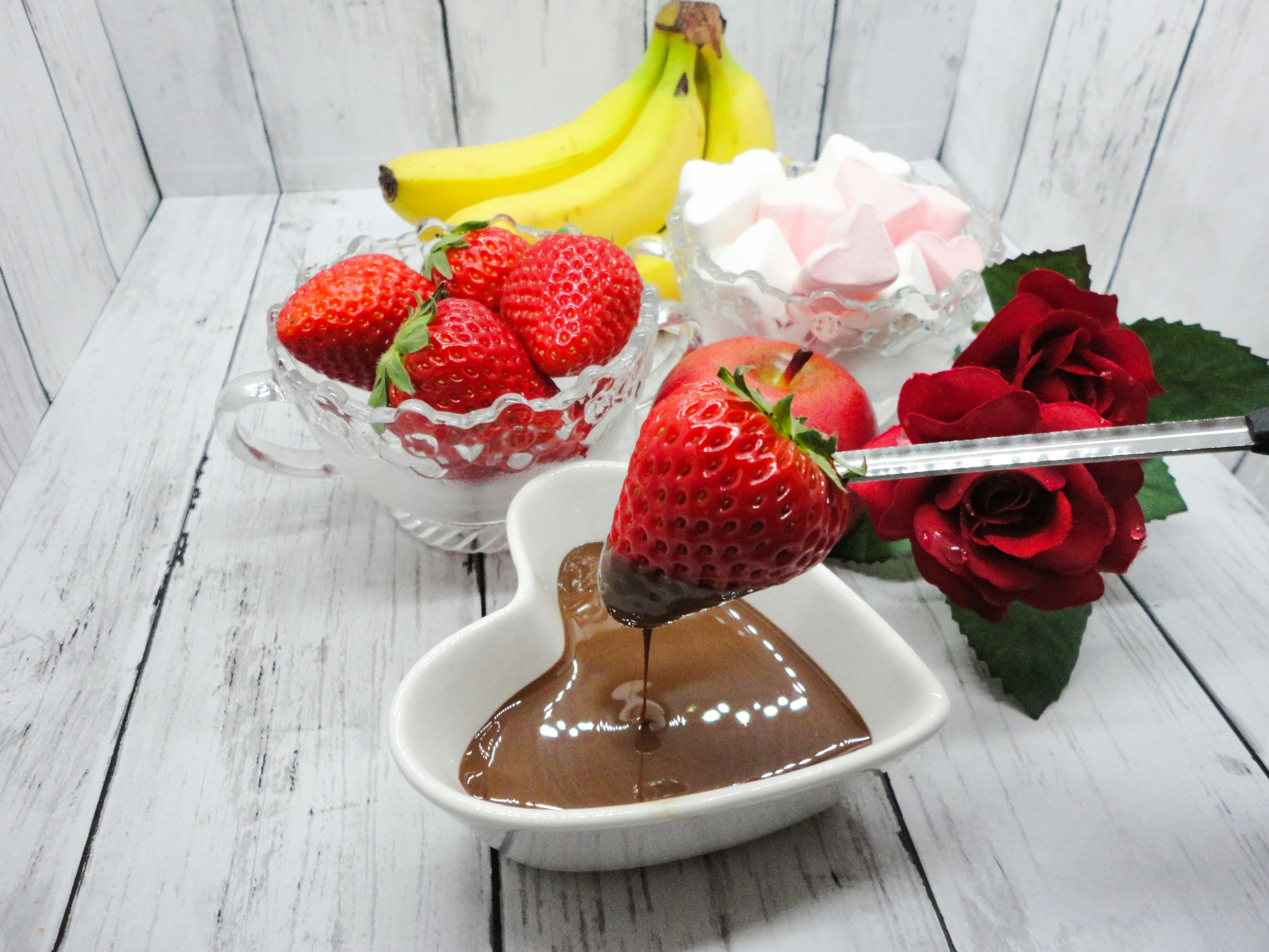 Un piatto a forma di cuore con salsa di cioccolato e una fragola immersa, circondata da banane e fiori