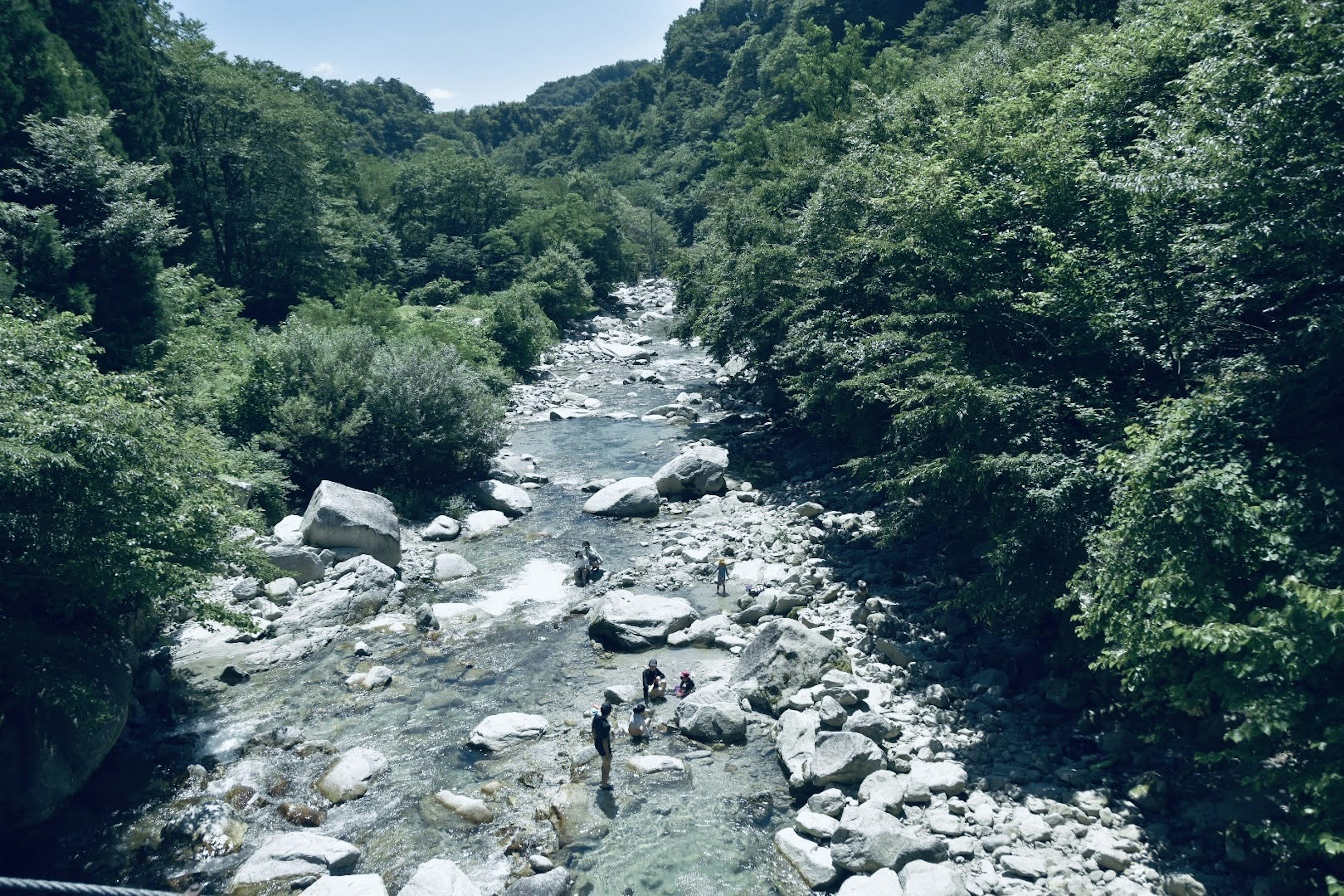 緑豊かな山間の川に石が散在し、二人の人が水辺で活動している風景