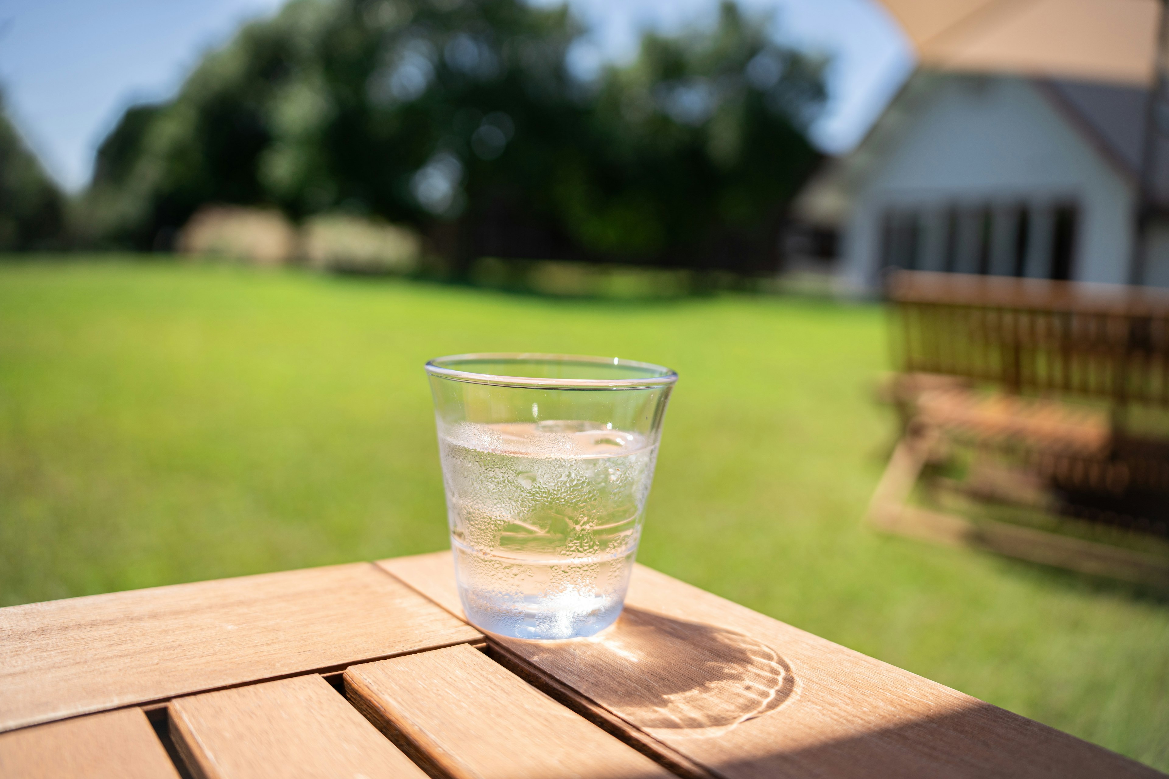 Un vaso transparente con agua helada colocado sobre una mesa de madera con césped verde y parte de una casa al fondo