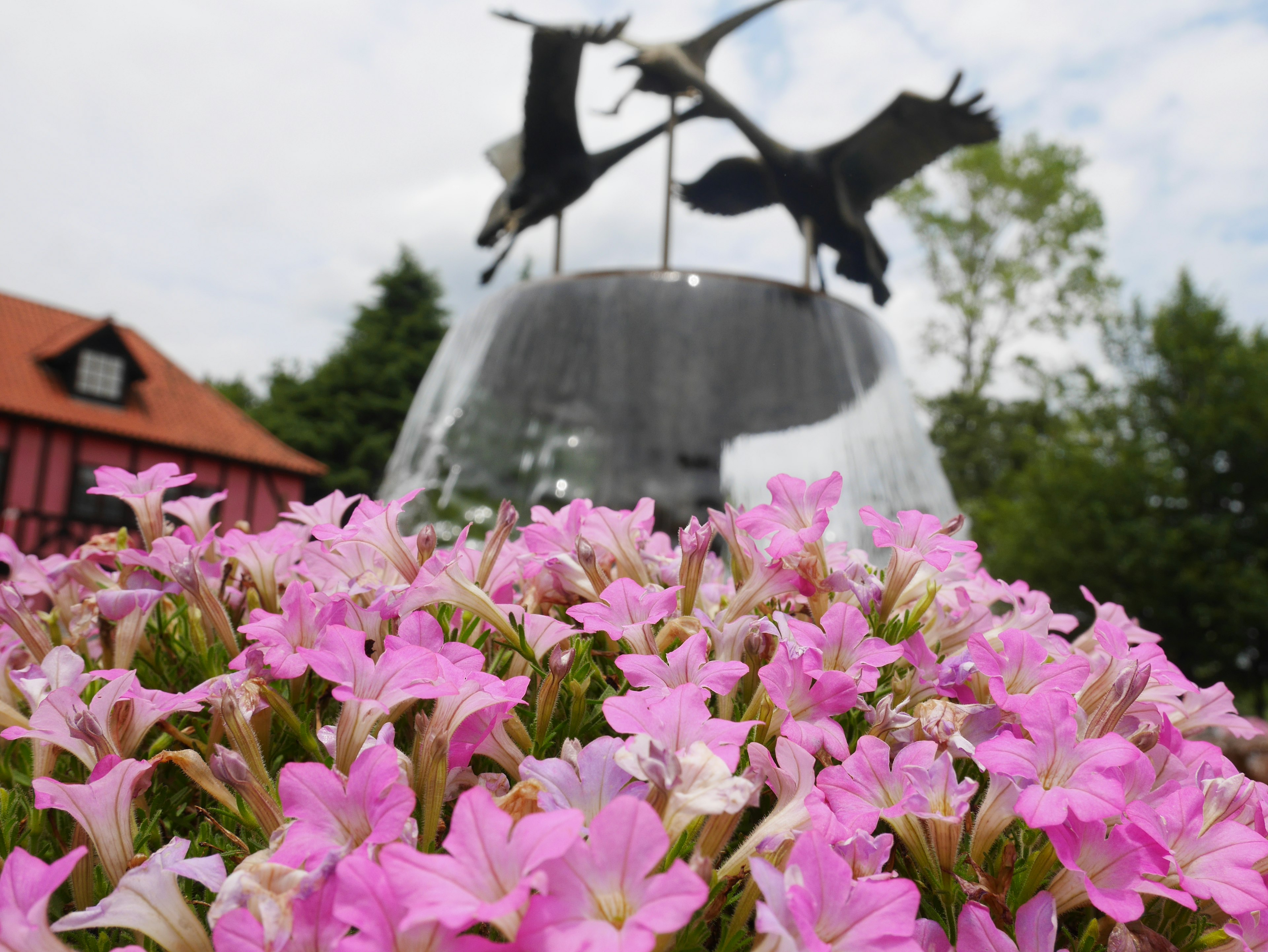 ピンクの花が咲いている前景に噴水の彫刻があり、背景に建物と木々が見える風景