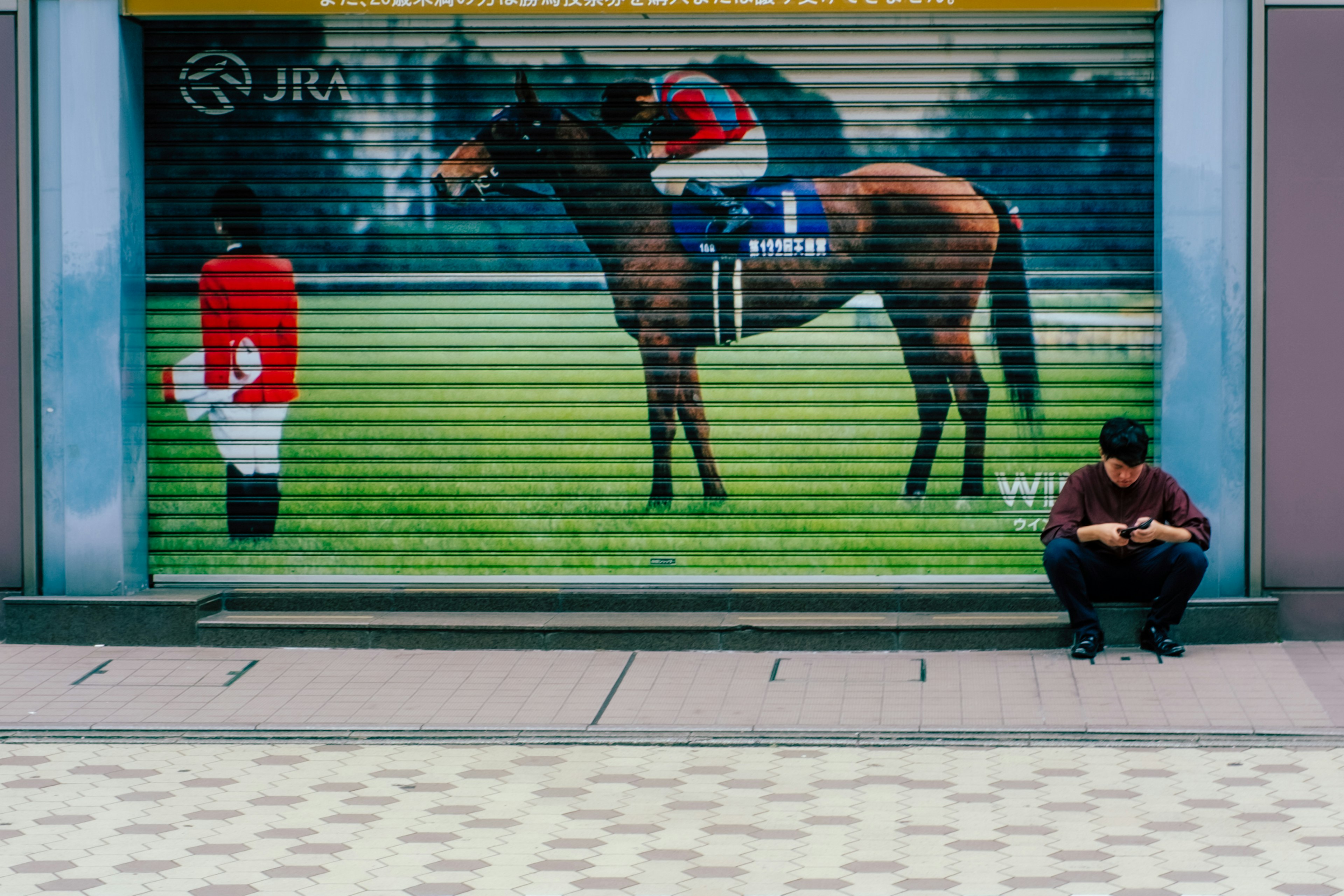 Ein Mann, der auf dem Boden sitzt und ein Smartphone benutzt, vor einem großen Wandgemälde eines Pferdes und eines Jockeys auf einem Rollladen