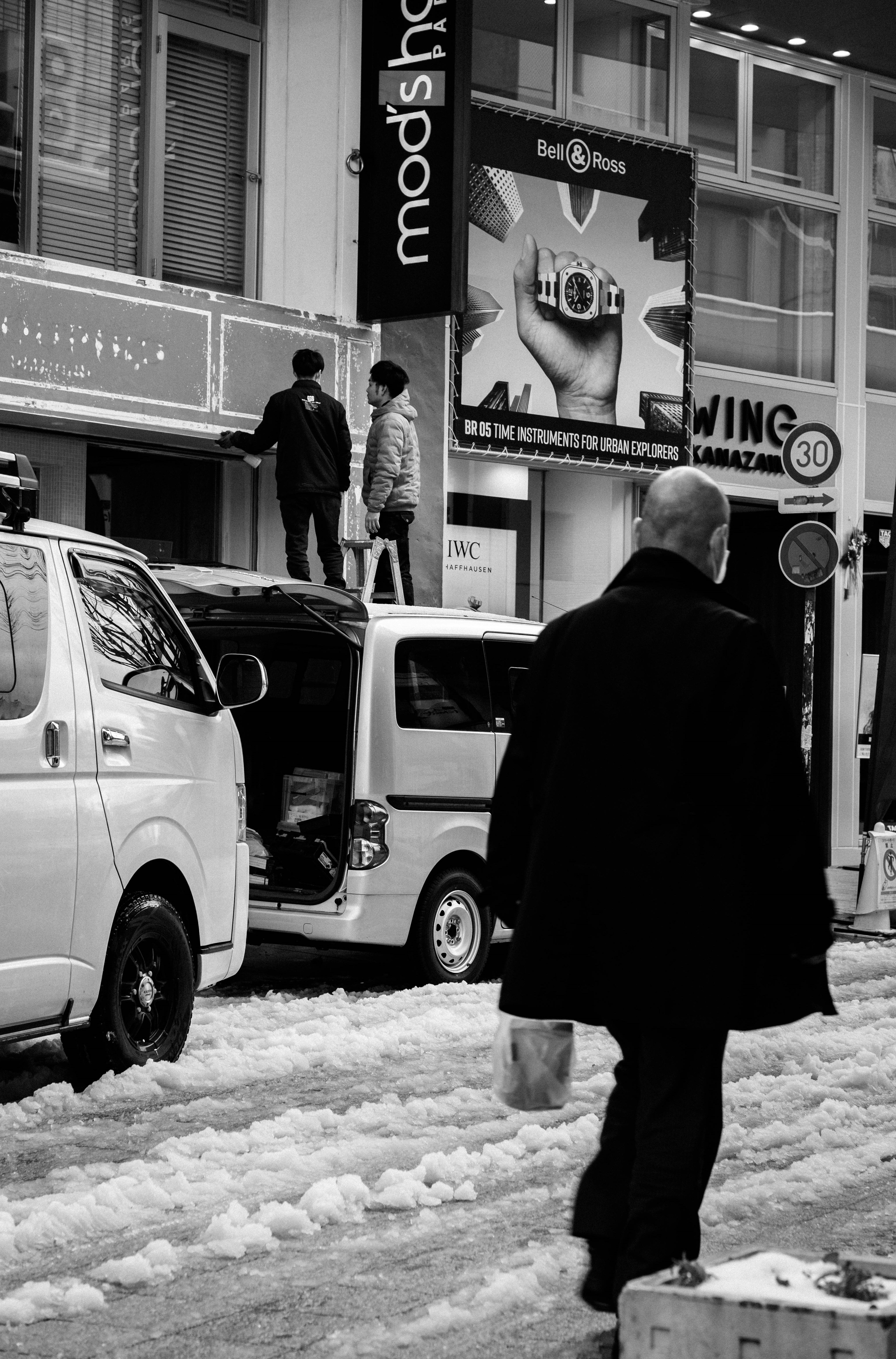 Foto en blanco y negro de un hombre caminando por una calle nevada con trabajadores arriba