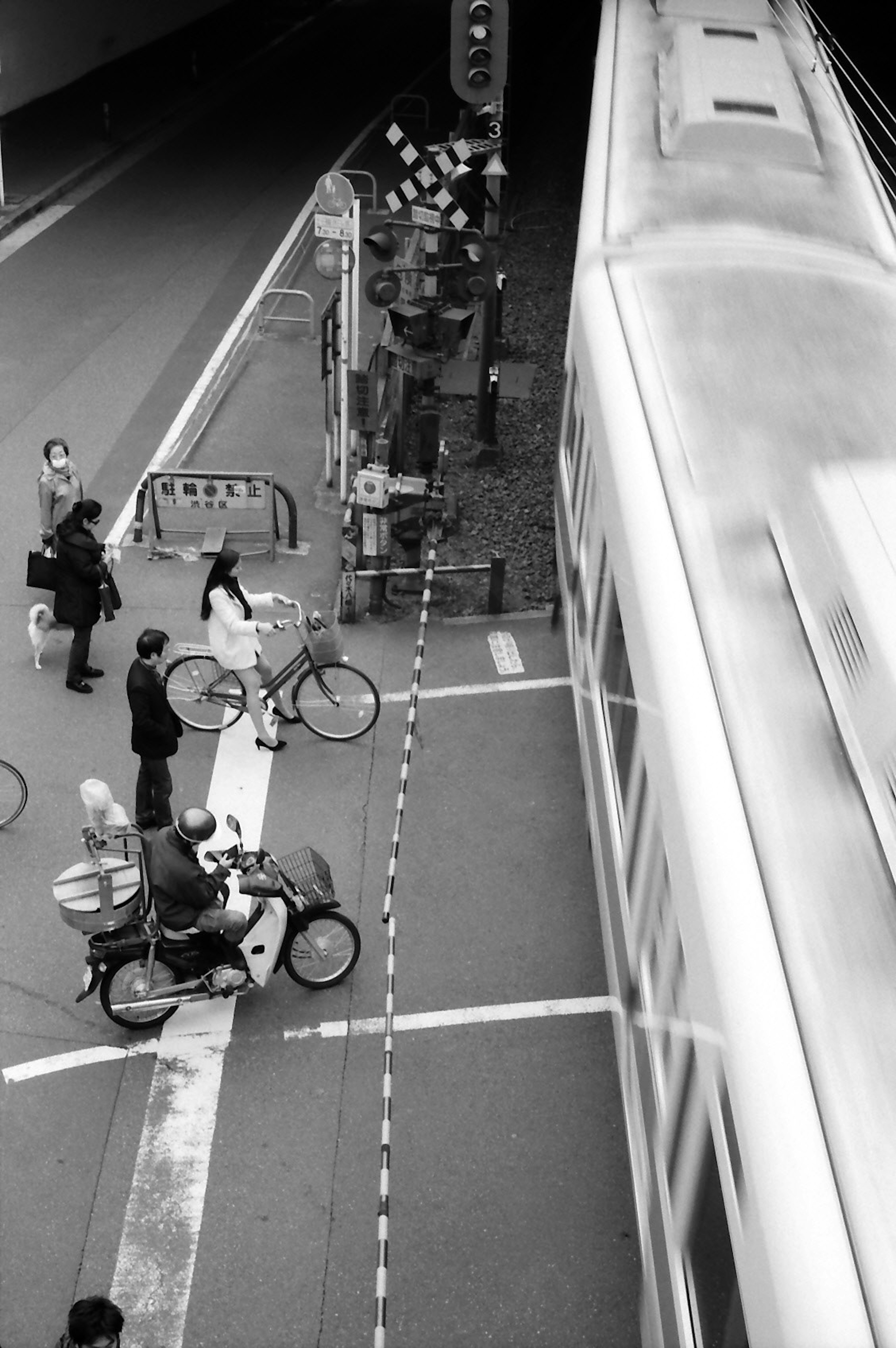 Escena en blanco y negro de ciclistas y peatones esperando en un paso de peatones cerca de una estación de tren