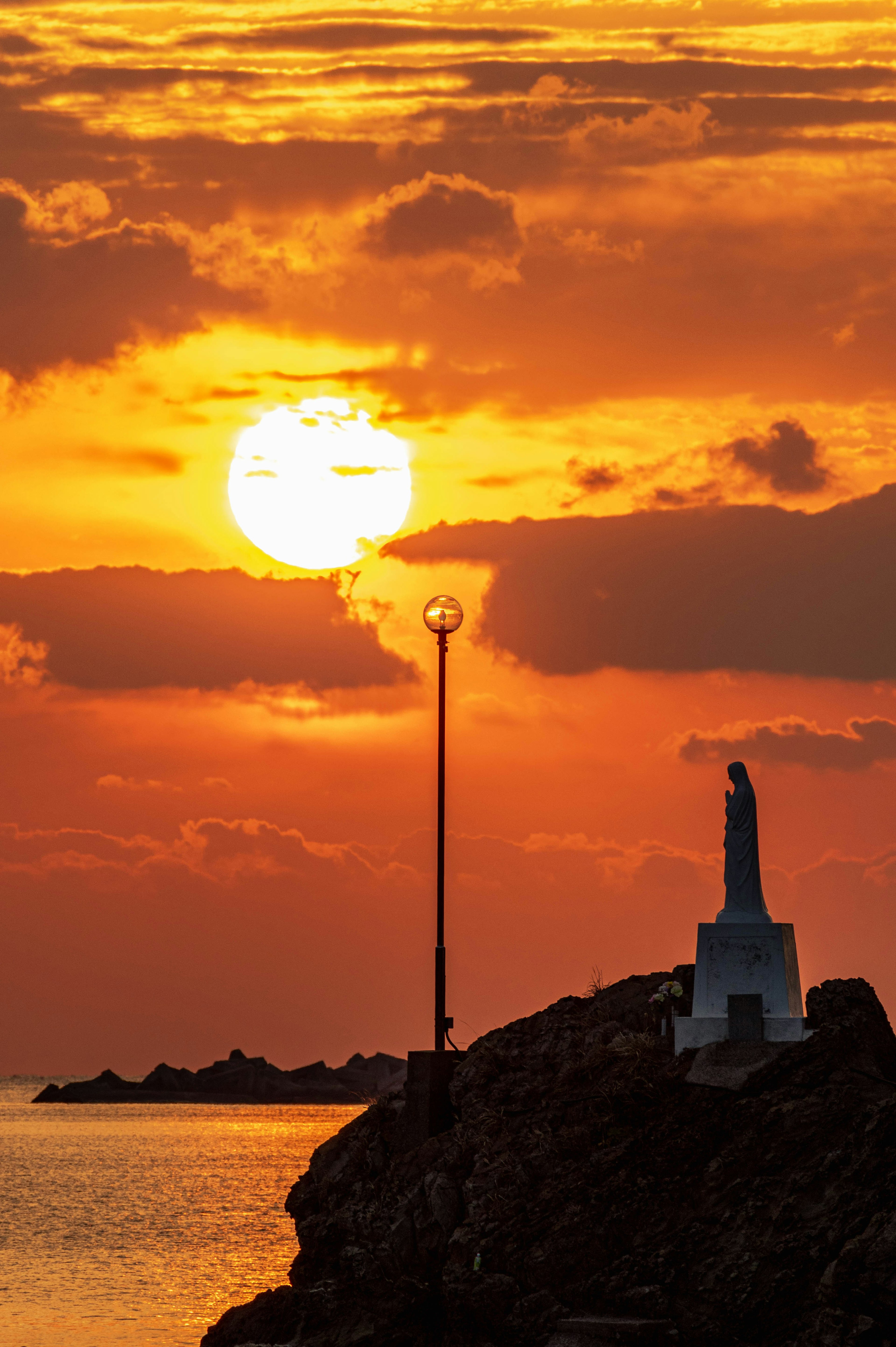 Silhouette of a statue and streetlight against a sunset over the ocean