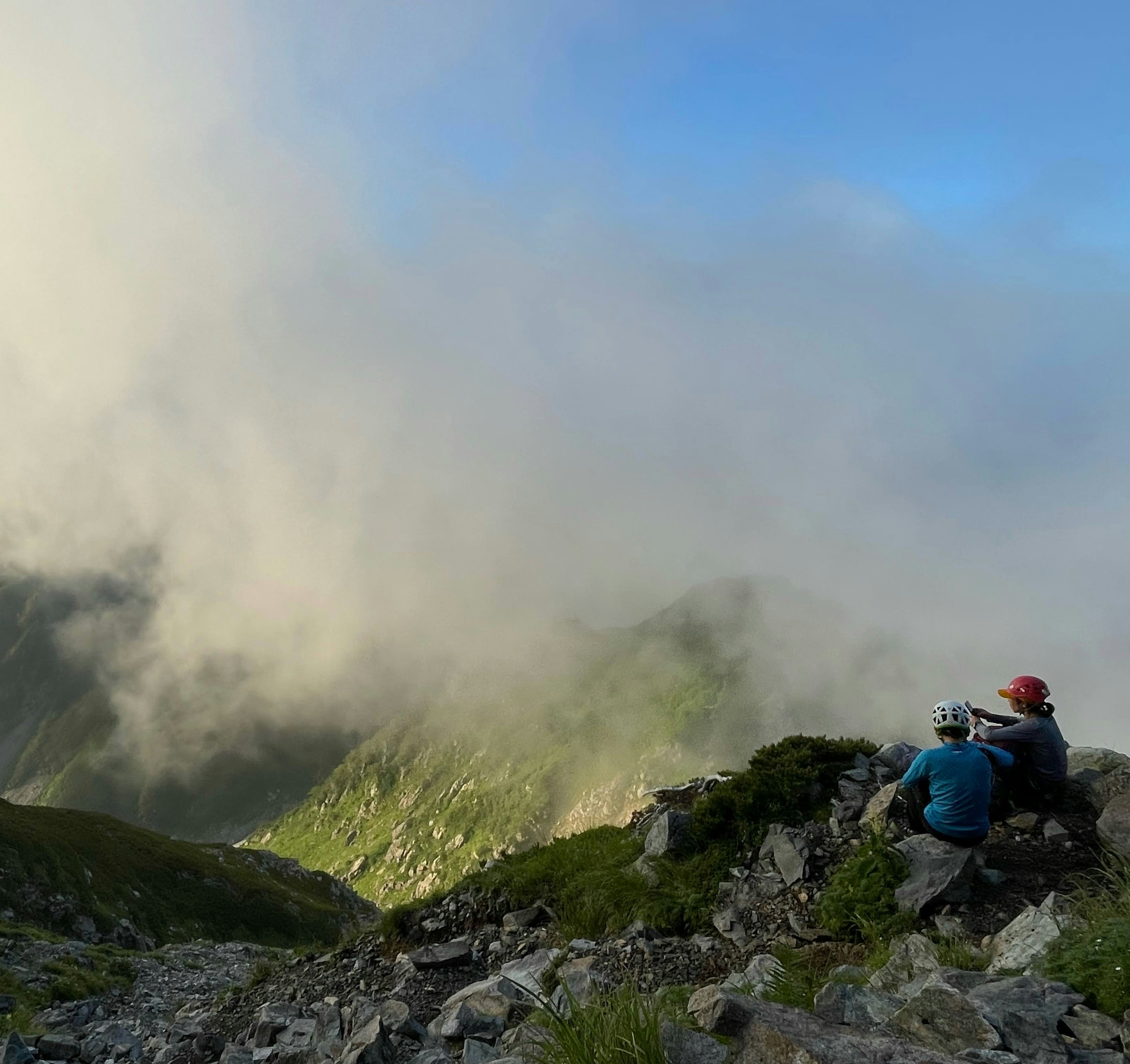 Dos escaladores sentados en una pendiente rocosa observando las montañas envueltas en niebla