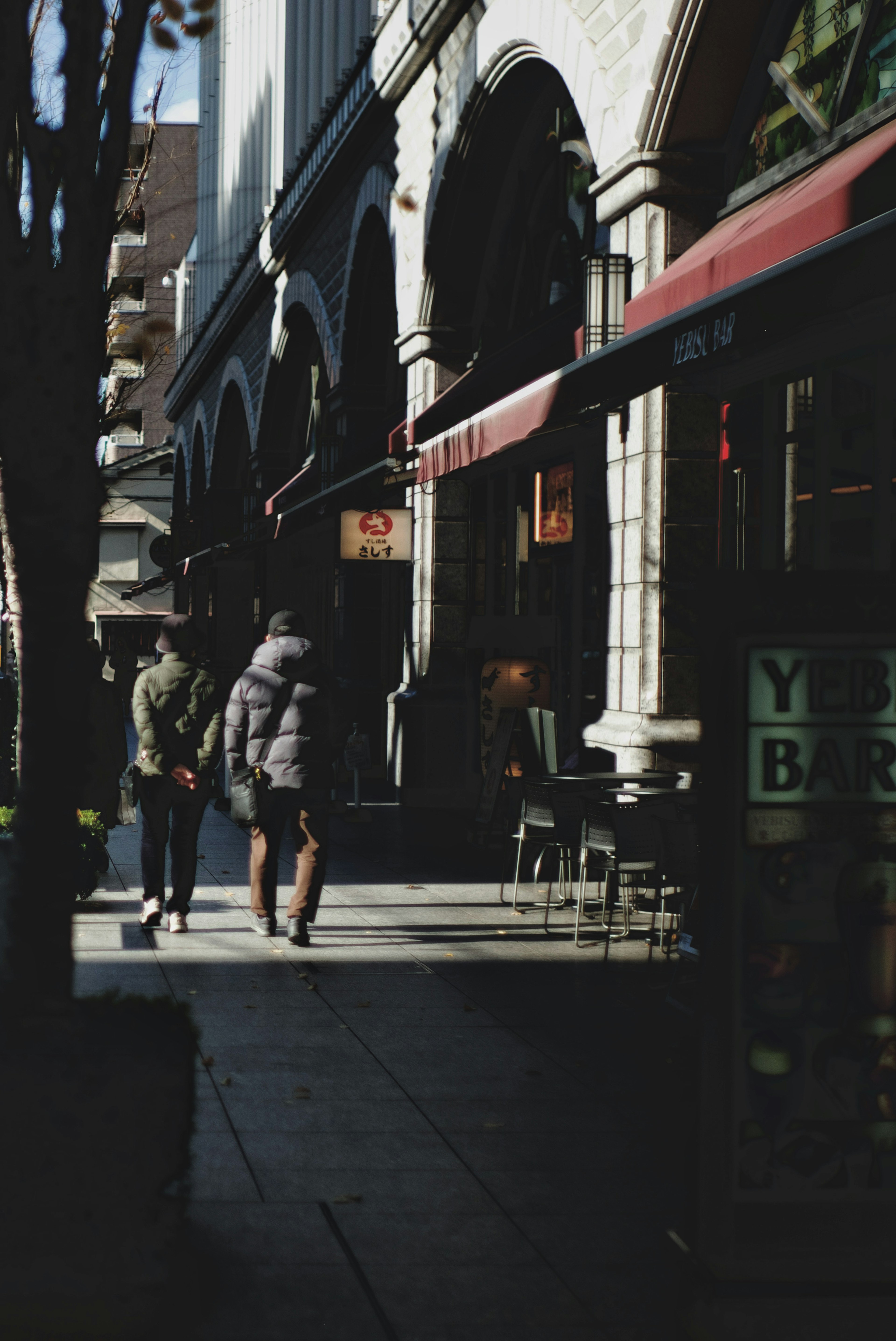 Two individuals walking in front of archway buildings and a café