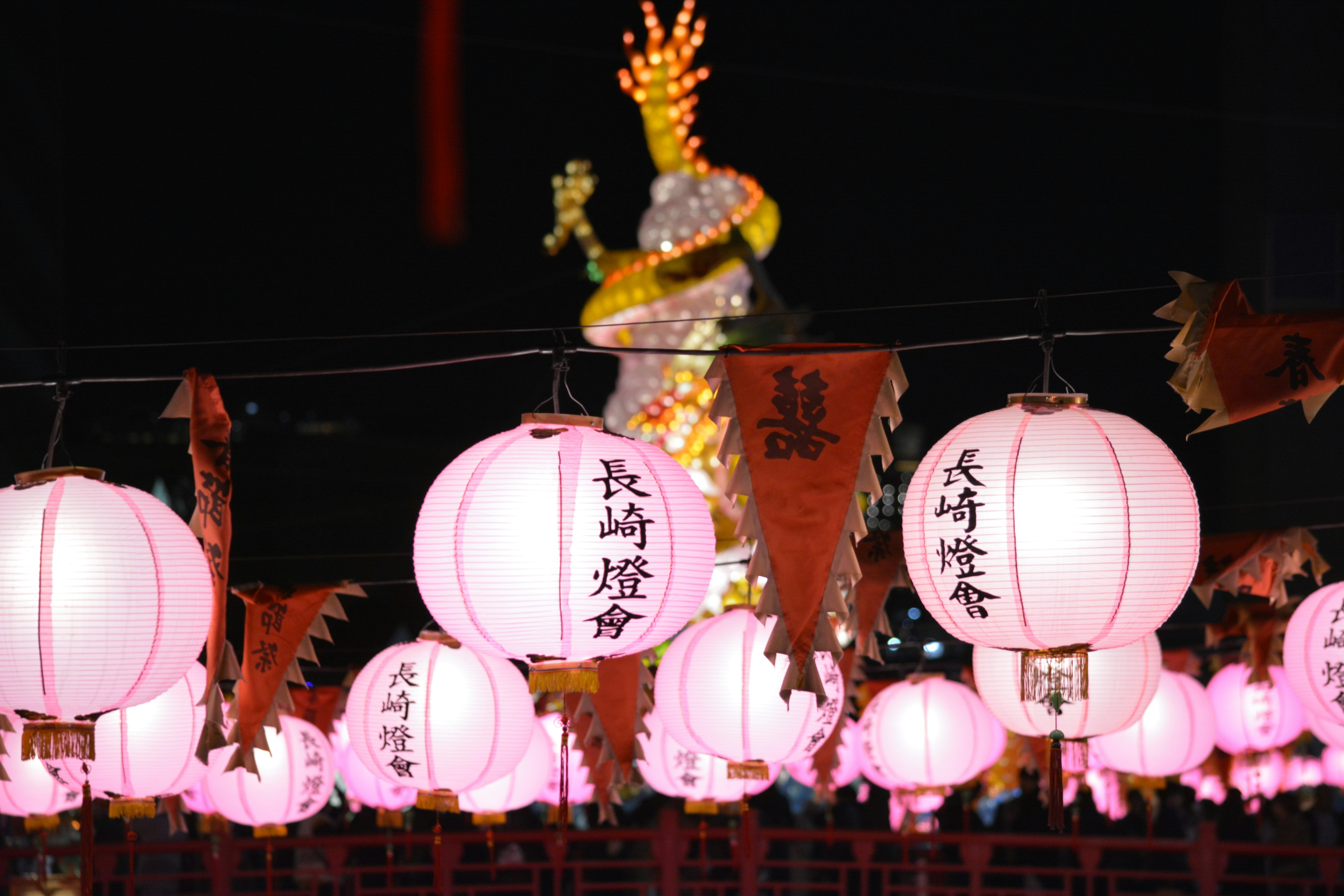 夜の祭りの風景でピンクの提灯が並び、後ろには明るい装飾が施された像が見える