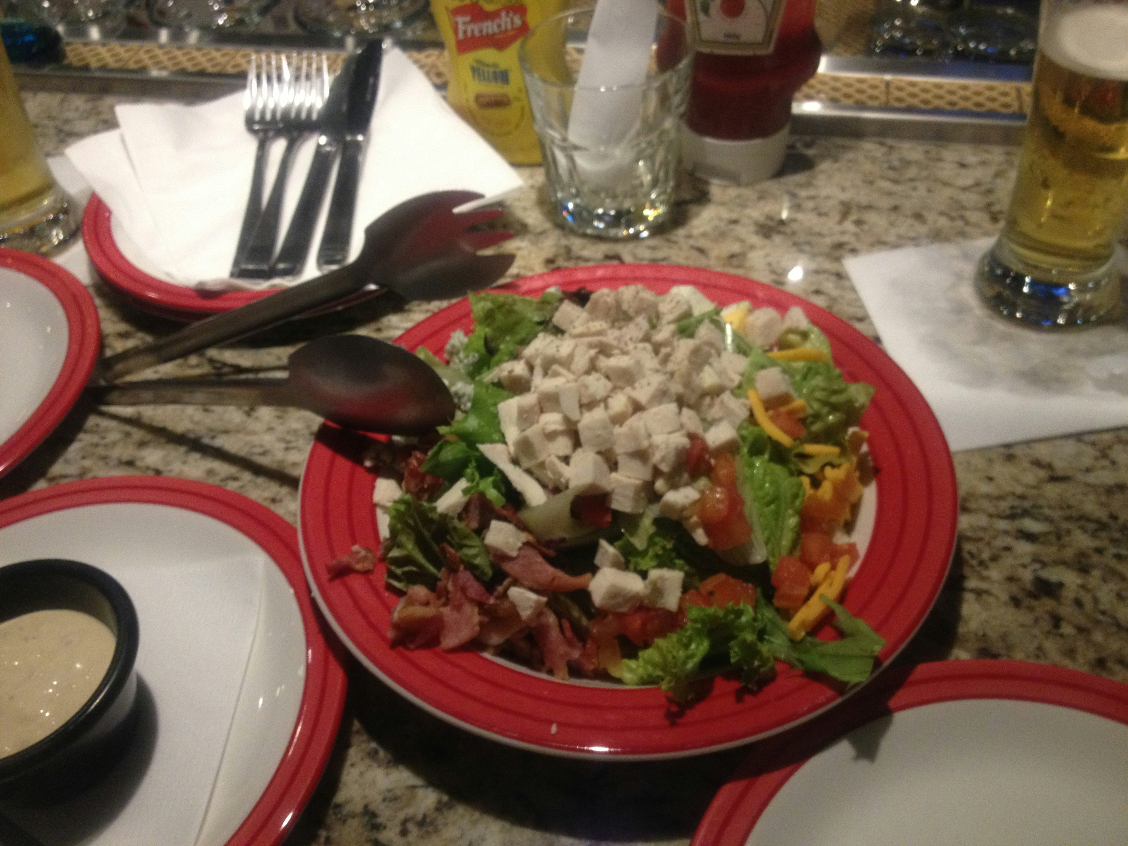 Una ensalada colorida con varios vegetales y pollo servida en un plato rojo con bebidas en la mesa