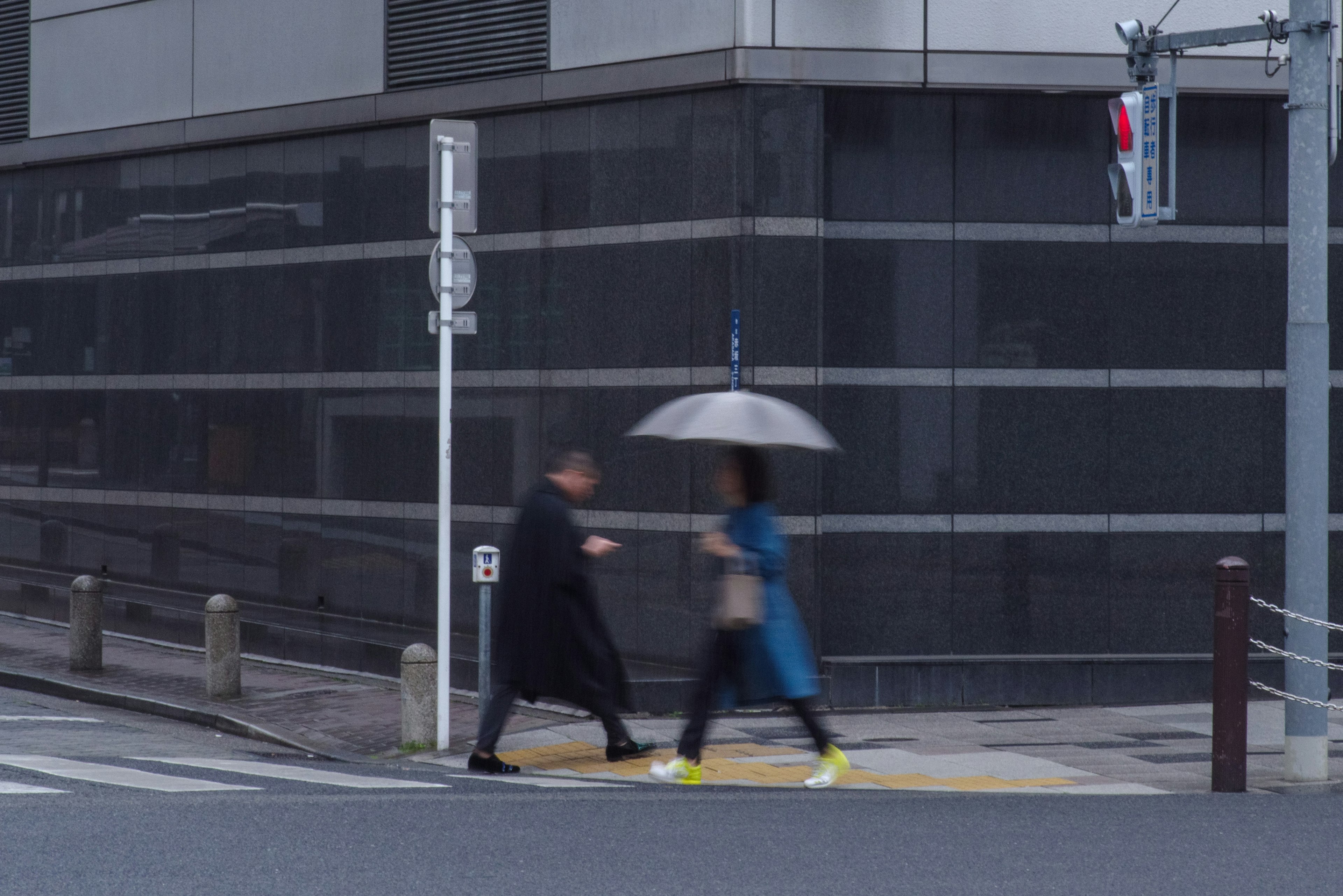 A woman with an umbrella and a man in a black coat crossing the street with a modern building in the background