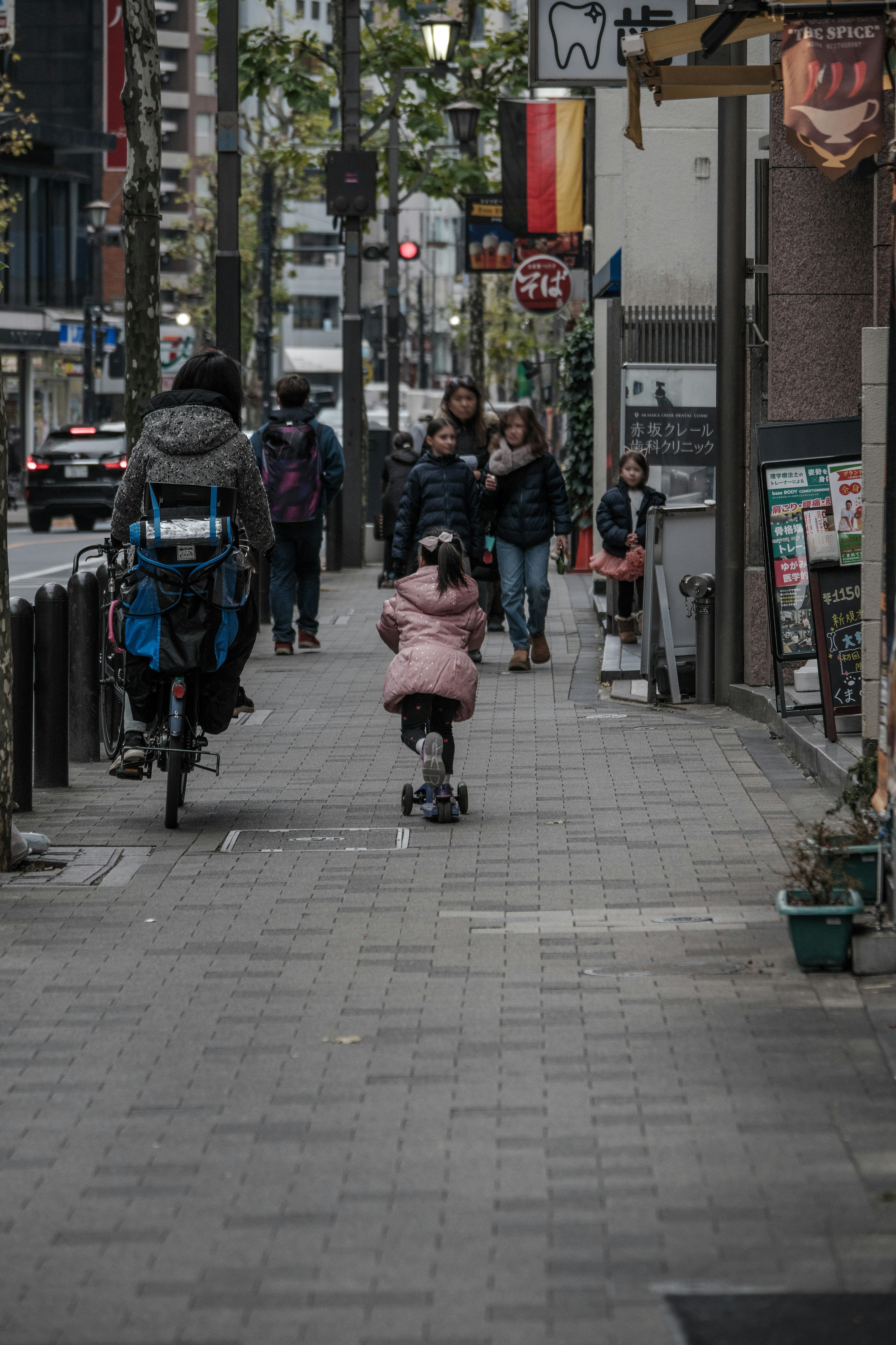 人々が歩いている通りにピンクのコートを着た子供がスクーターに乗っている