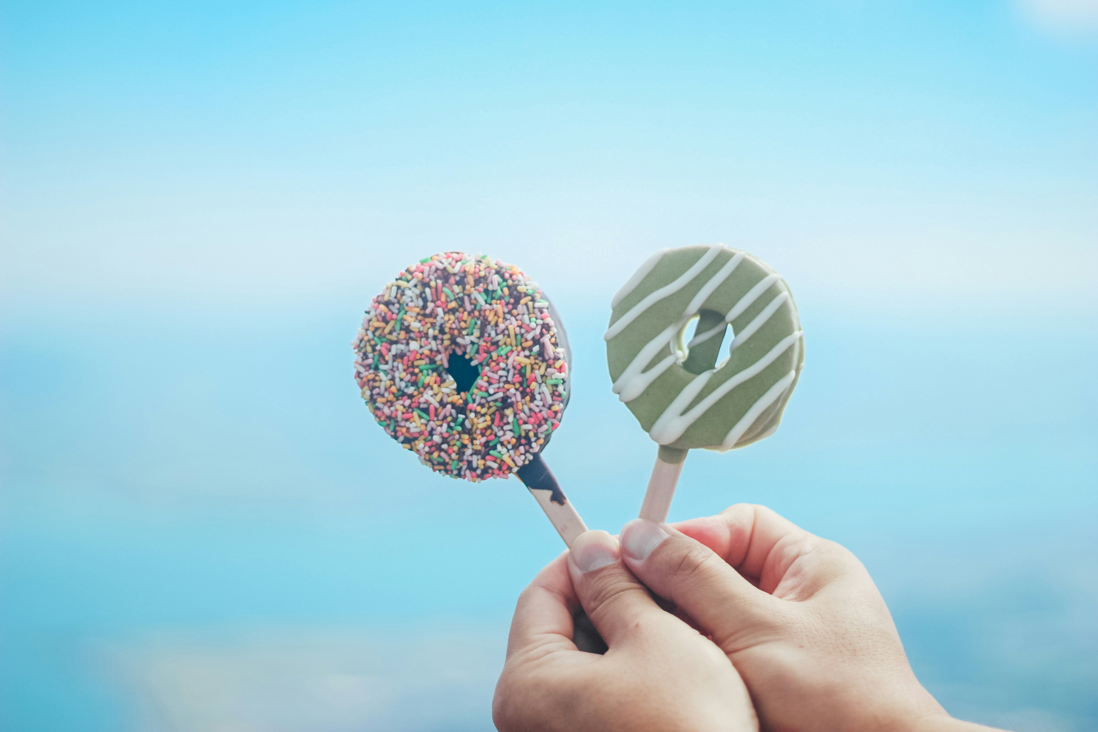 Due ghiaccioli a forma di donut colorati tenuti in mano contro un cielo blu