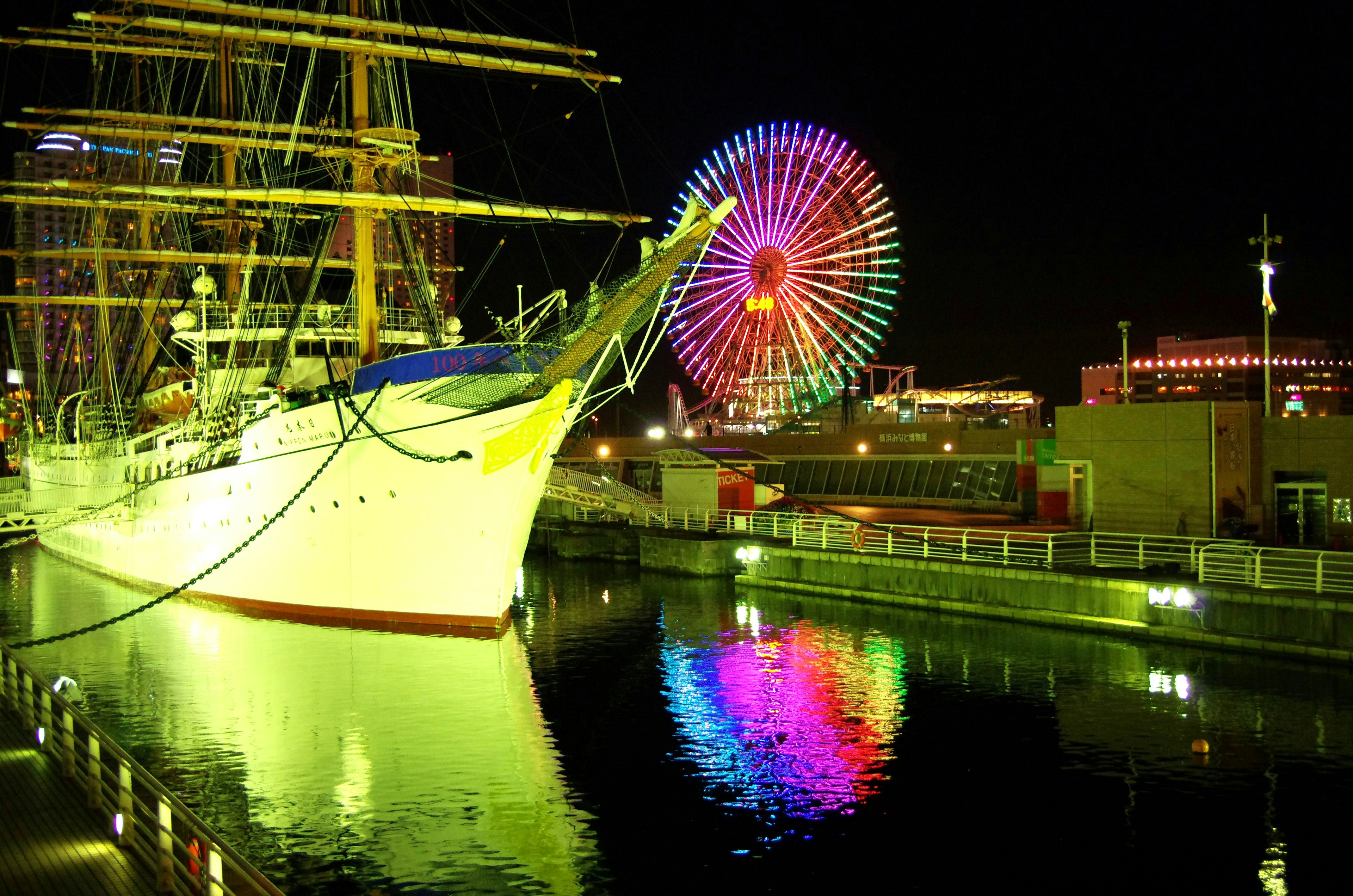 Una bella nave a vela illuminata di notte accanto a una ruota panoramica colorata che si riflette nell'acqua