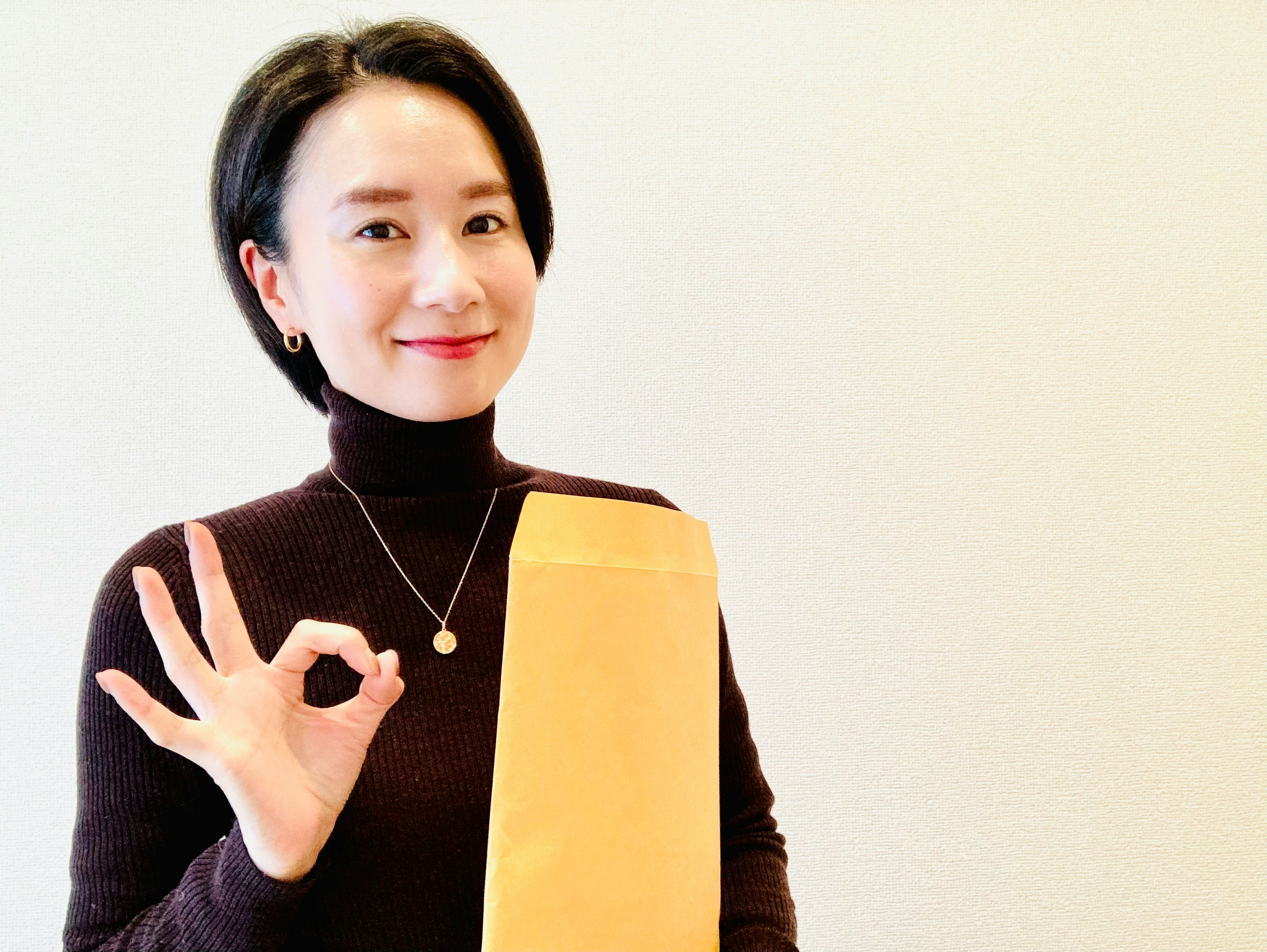 A woman in a brown turtleneck holding an orange envelope making an OK sign with her hand