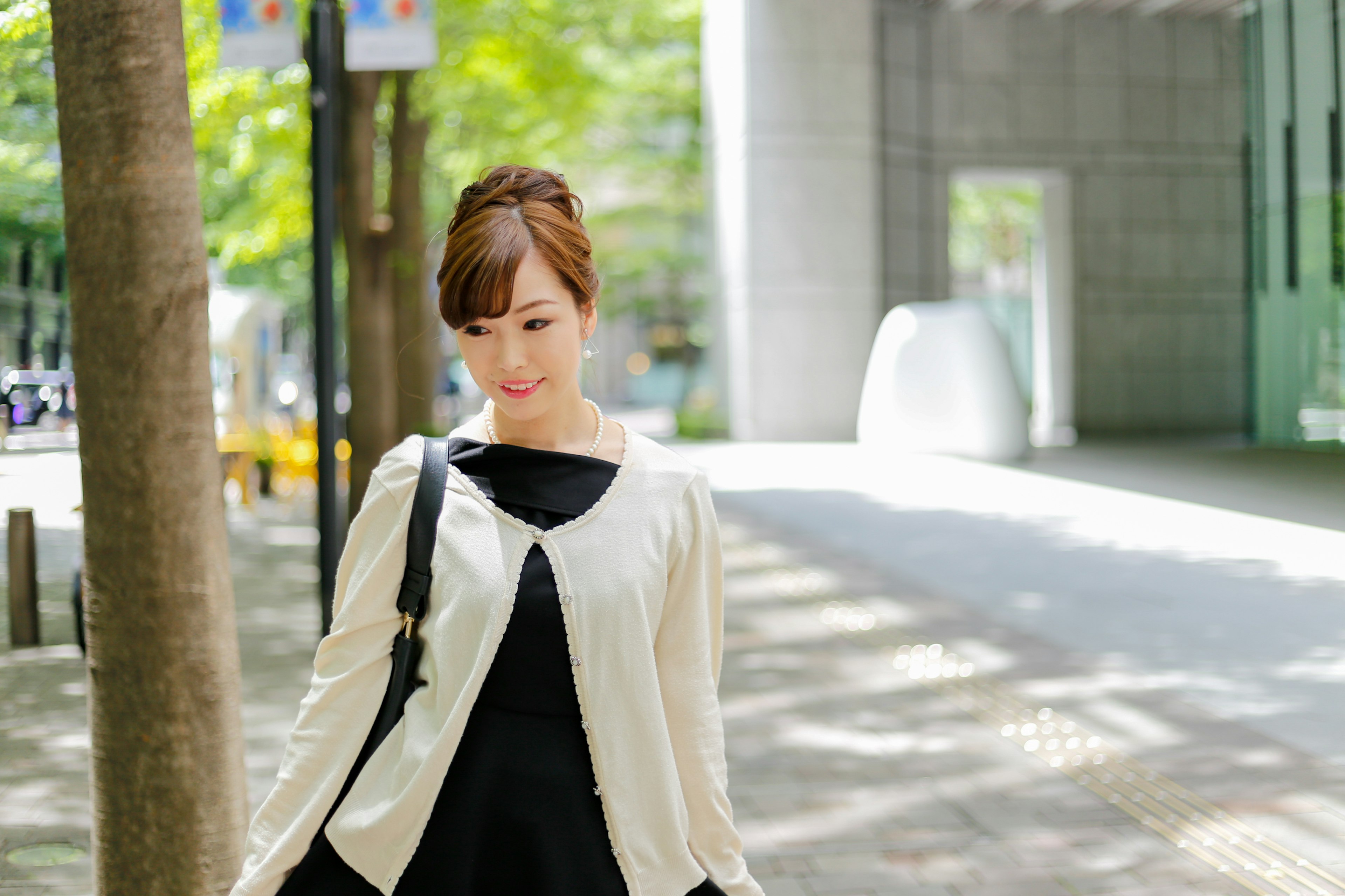 Woman walking on a park path wearing a white cardigan and black dress with green trees and modern buildings in the background