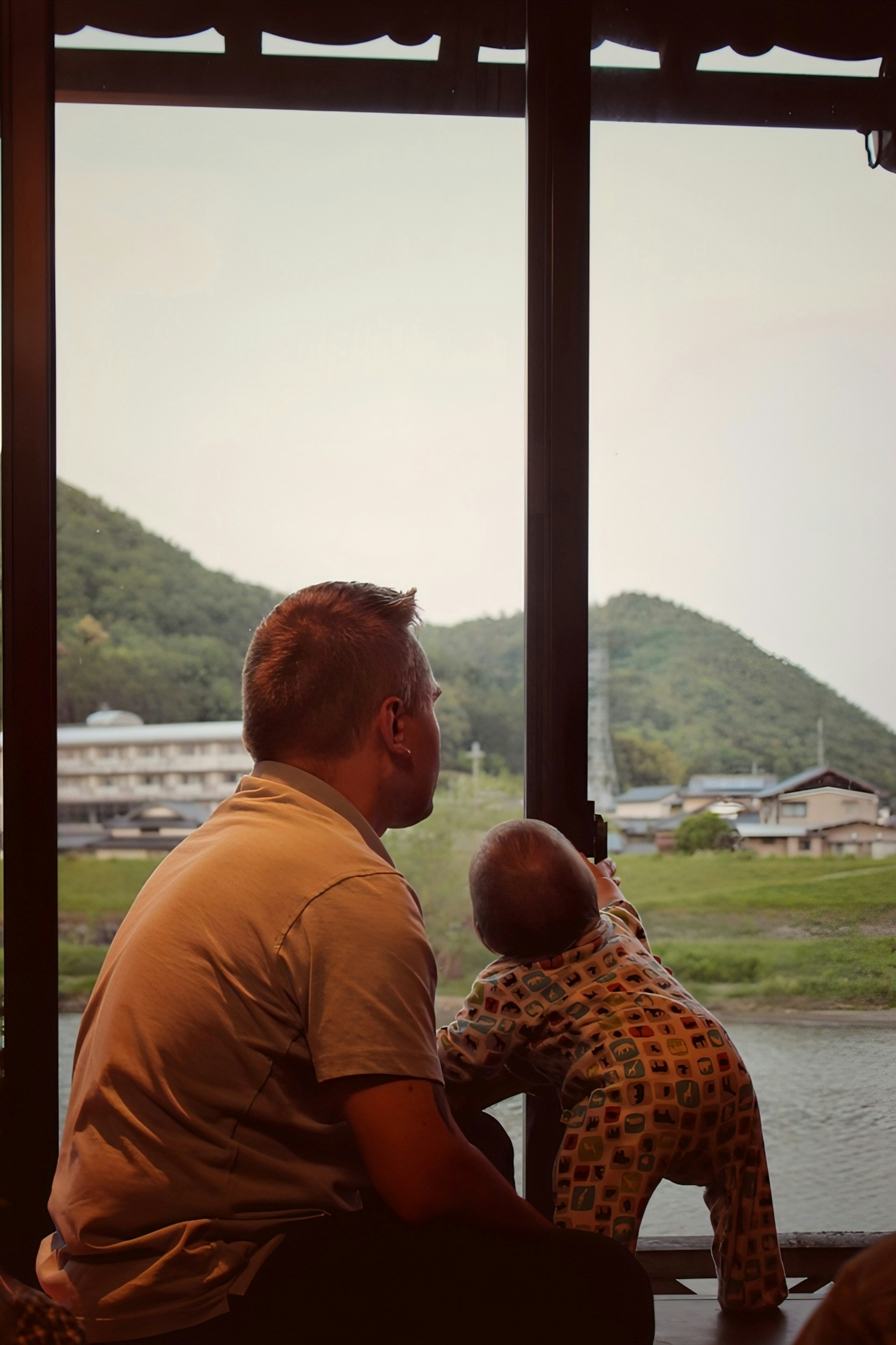 Padre y bebé mirando por la ventana a un paisaje escénico de montañas verdes y un río