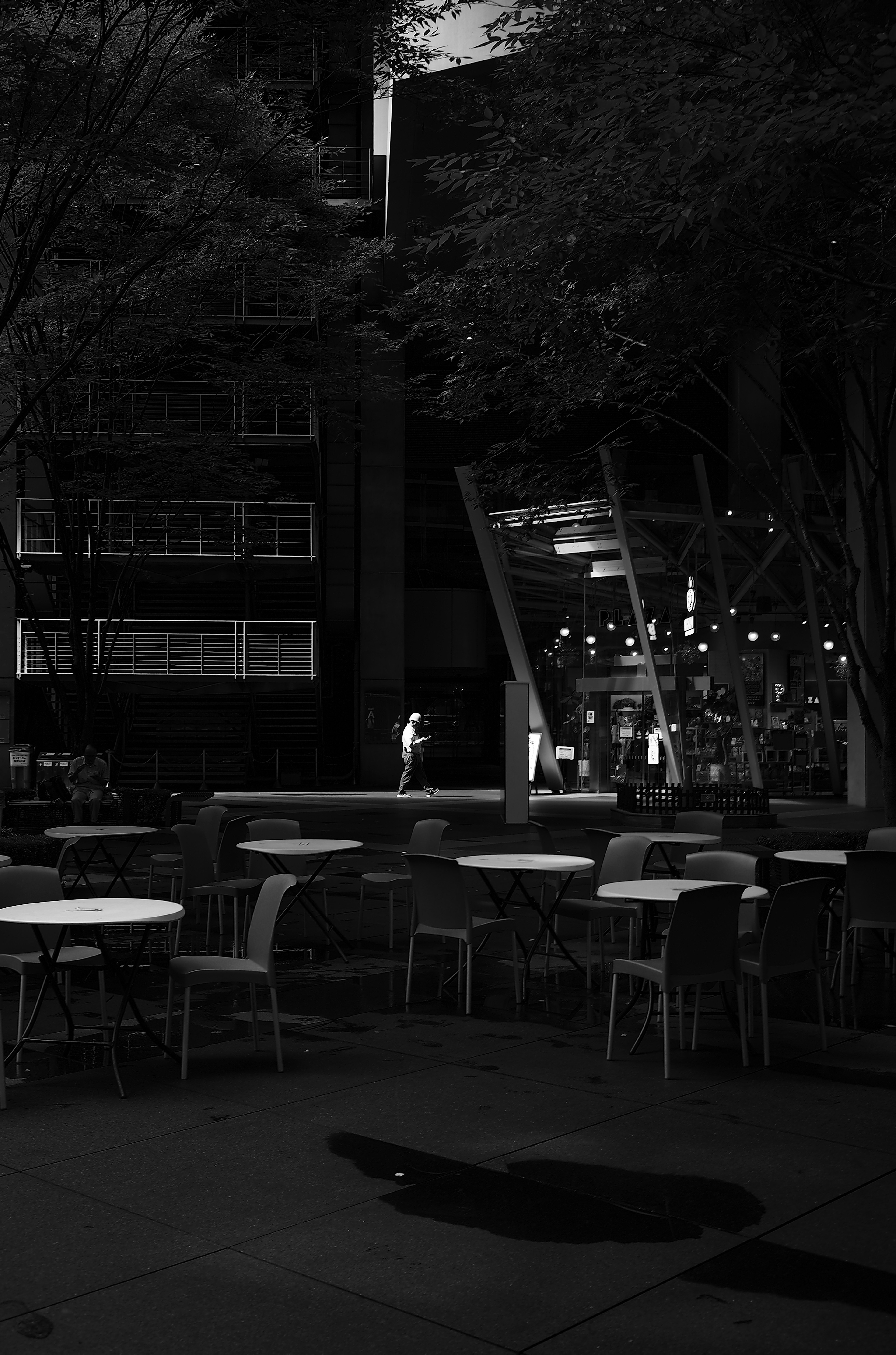 Foto en blanco y negro de una terraza de café nocturna con mesas y sillas rodeadas de árboles