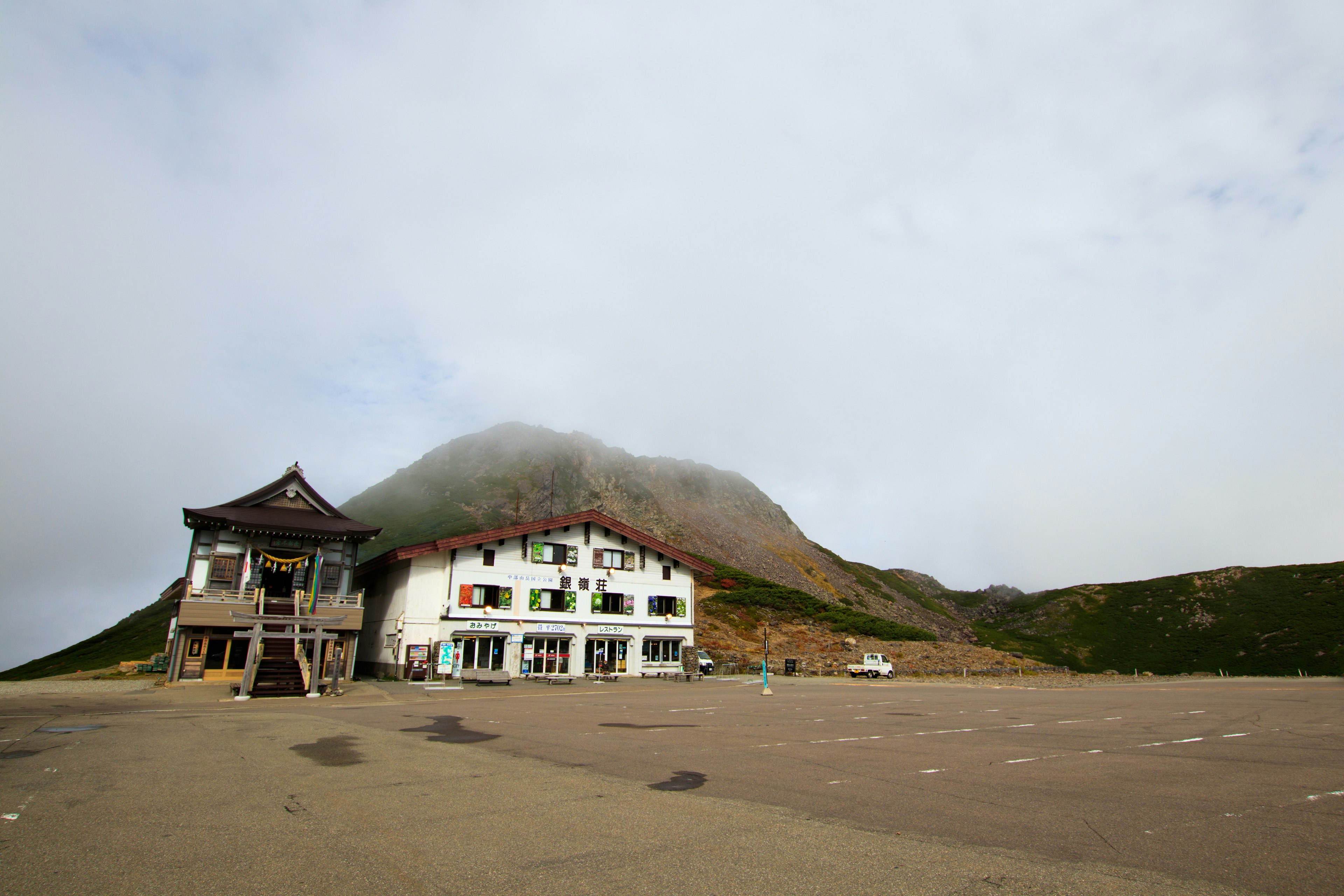 Two buildings near a mountain covered in clouds and a large parking area