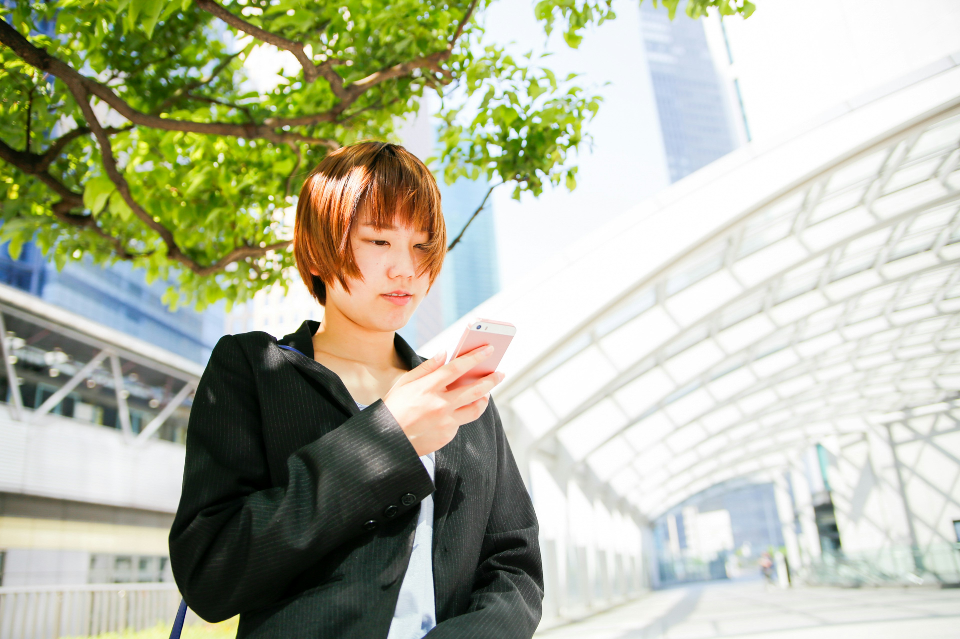 Junge Frau schaut auf ihr Handy unter einem grünen Baum mit moderner Architektur im Hintergrund