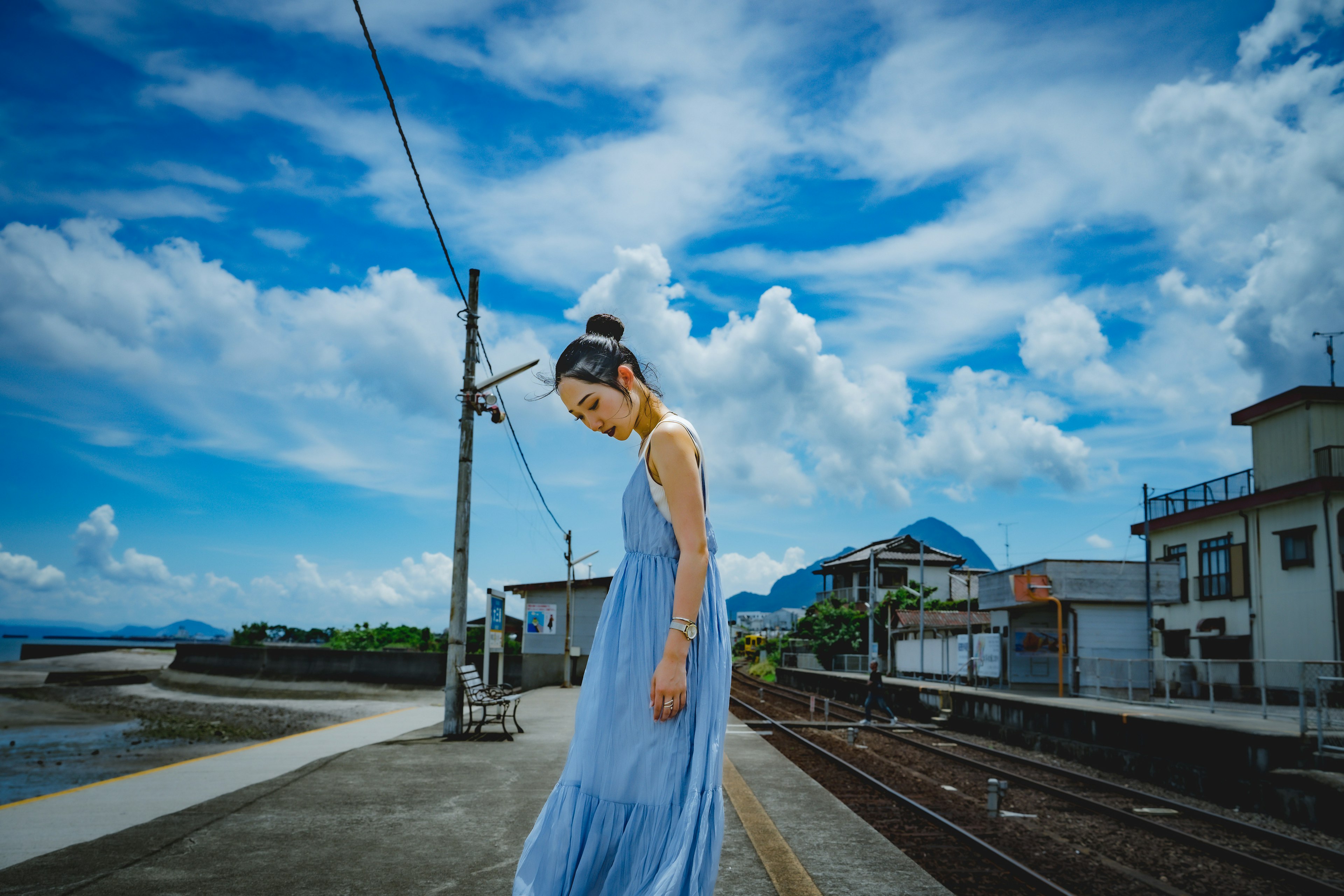 Una mujer con un vestido azul caminando cerca de las vías del tren bajo un cielo azul