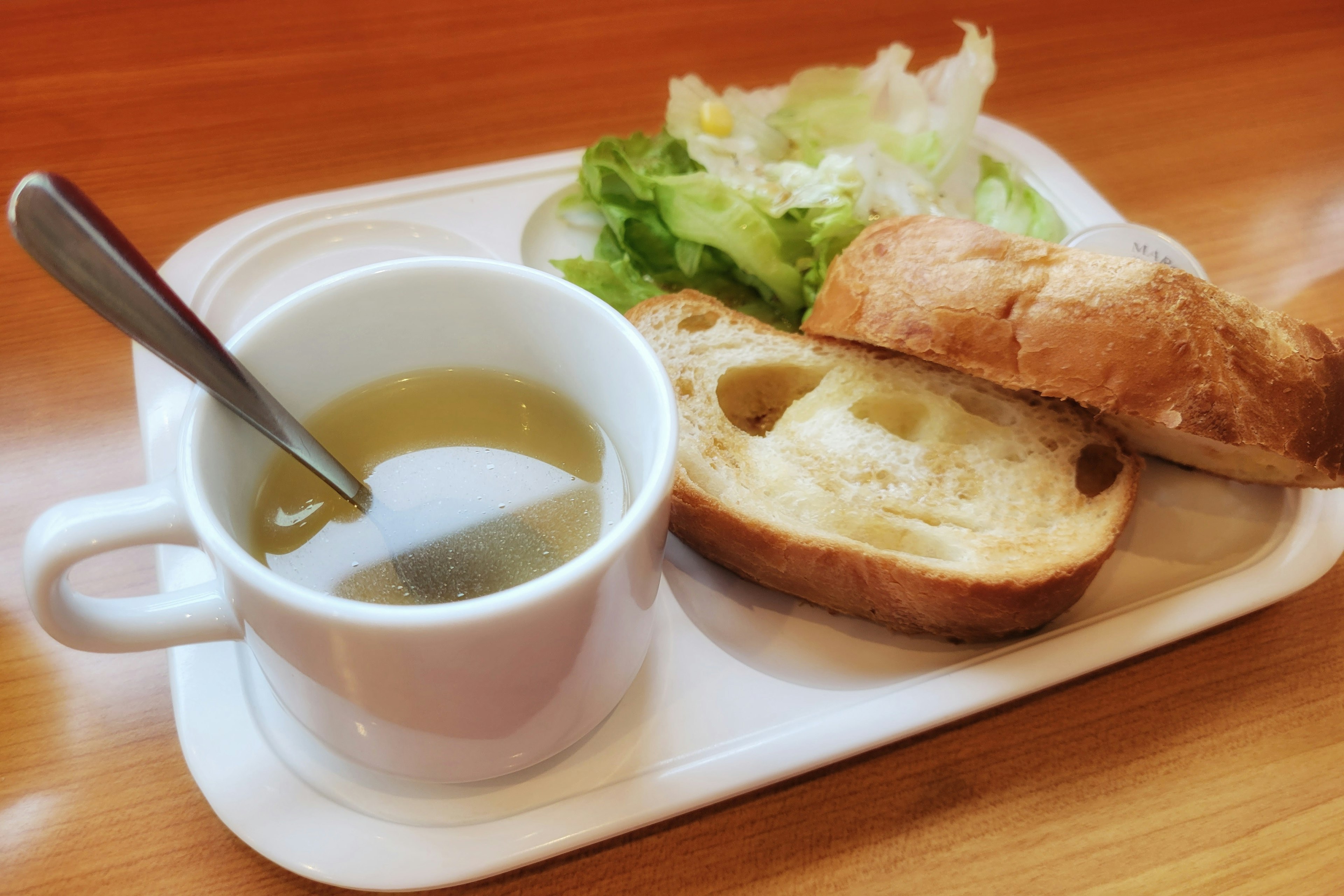 Weiße Tasse mit Suppe und Löffel, begleitet von Salat und Brot auf einem Teller