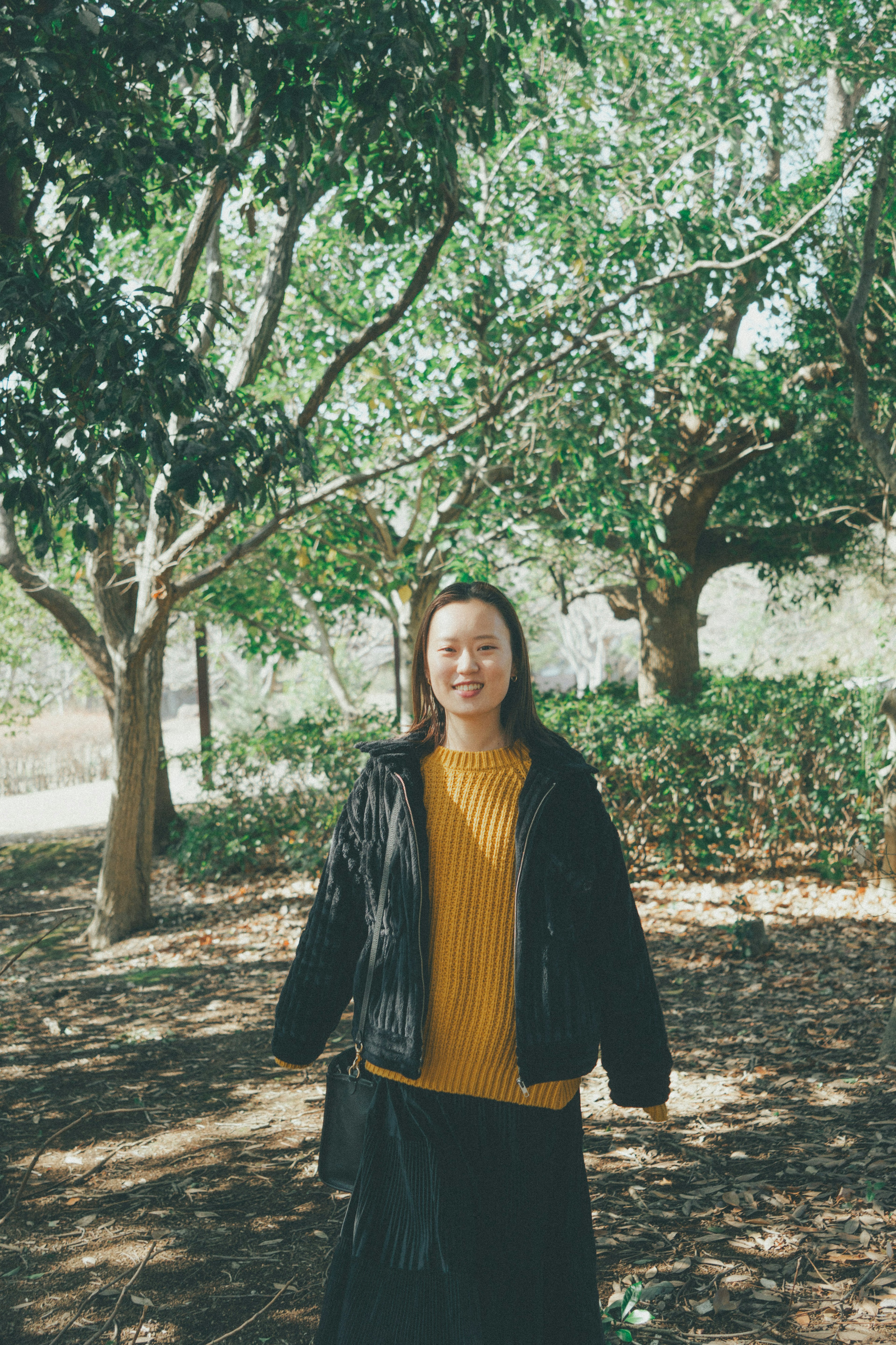 Femme se tenant parmi des arbres verts portant une veste noire et un pull jaune souriant dans la nature