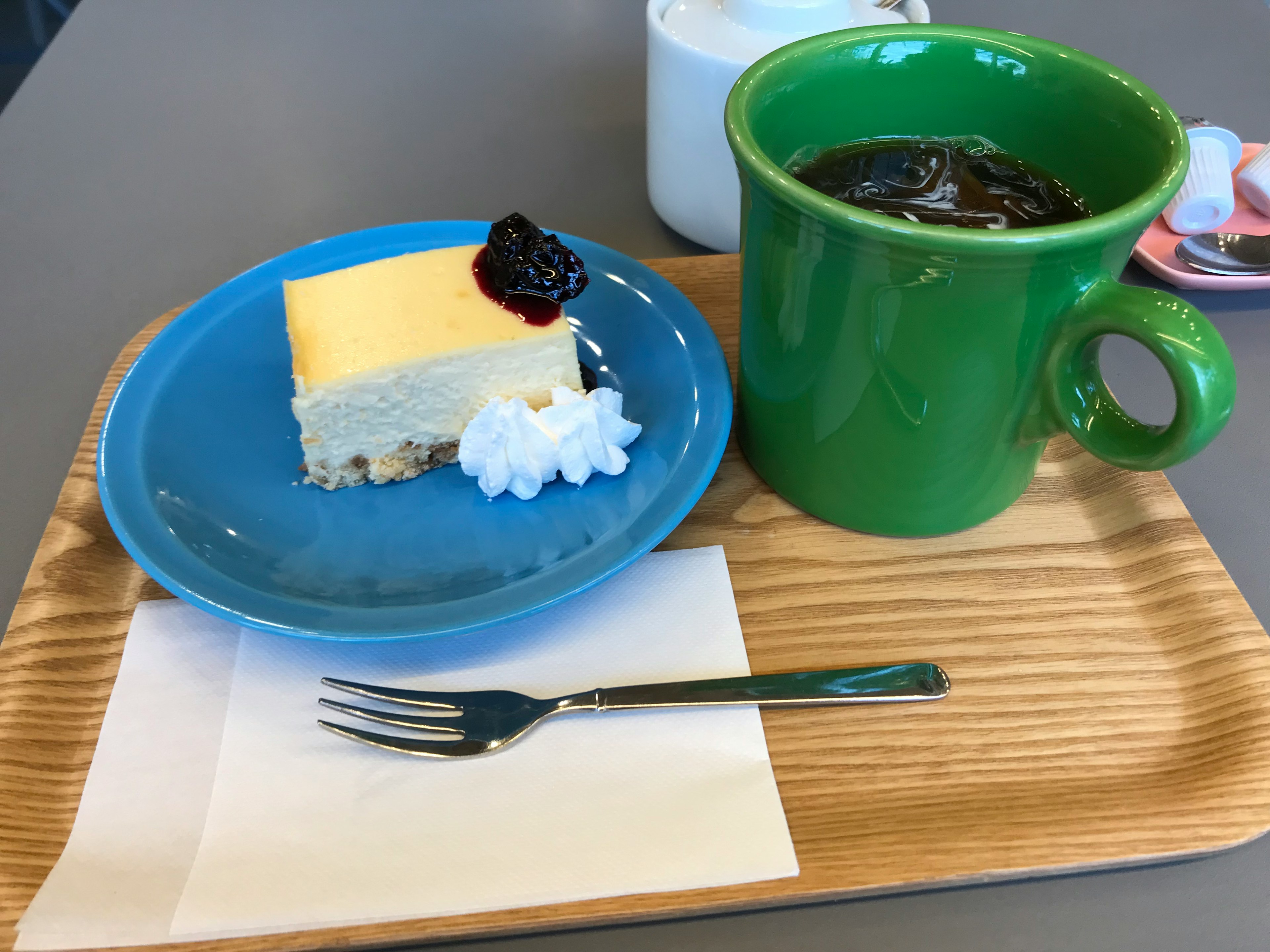 Ein Stück gelber Käsekuchen mit Schlagsahne und eine grüne Kaffeetasse auf einem Holztablett