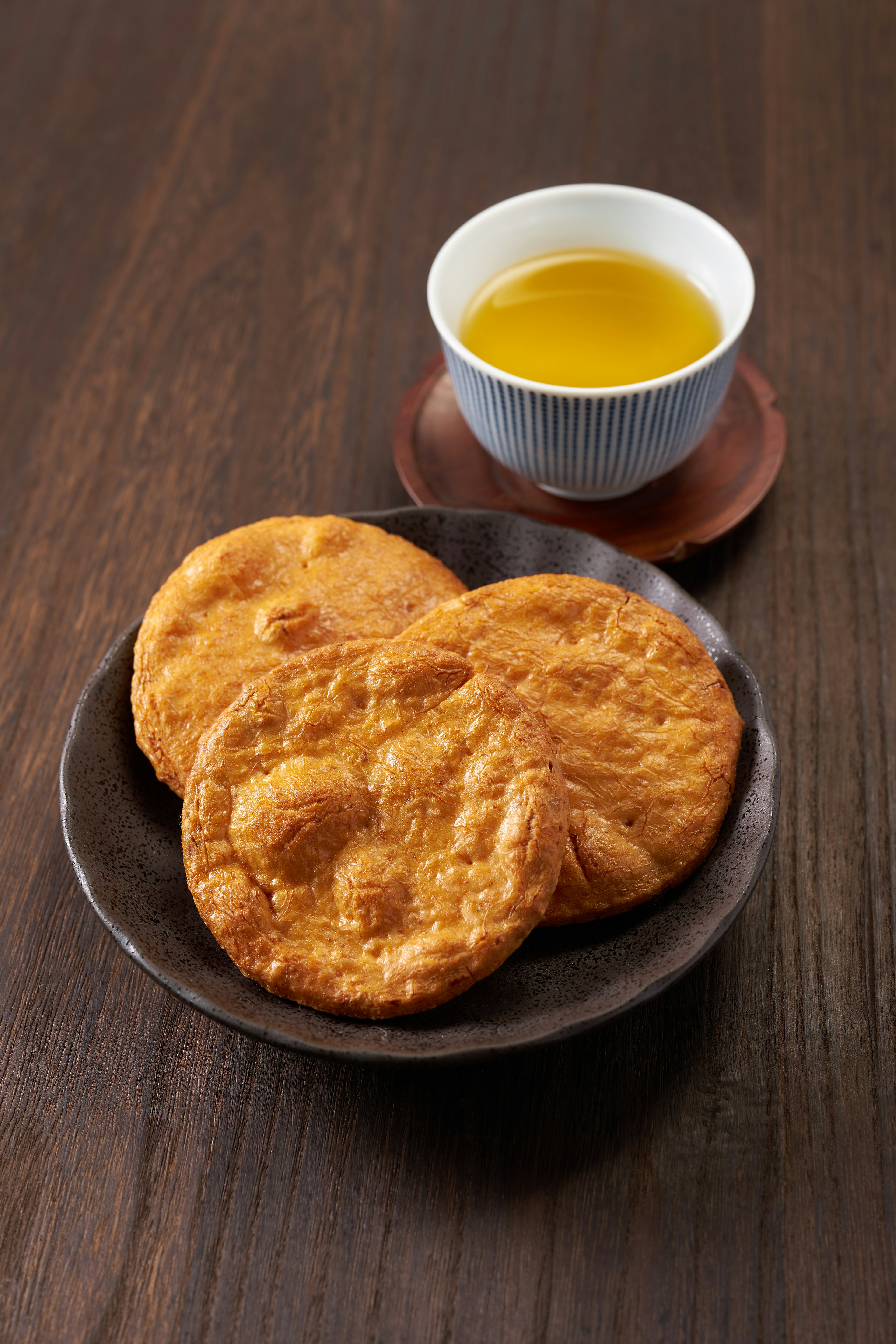 Three freshly baked cookies on a dark plate next to a cup of green tea on a wooden table