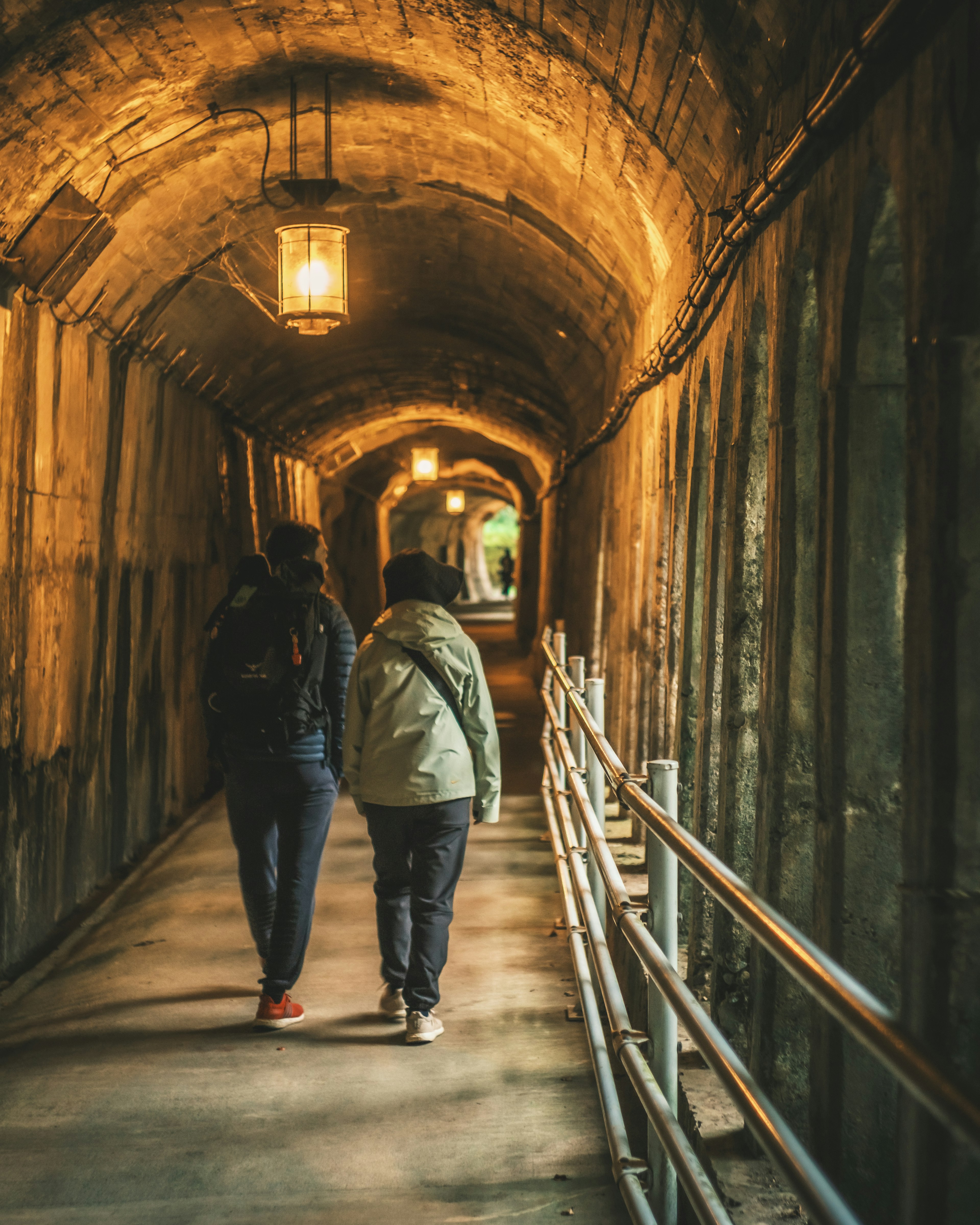 Two people walking in a dimly lit tunnel with ceiling lights and stone walls