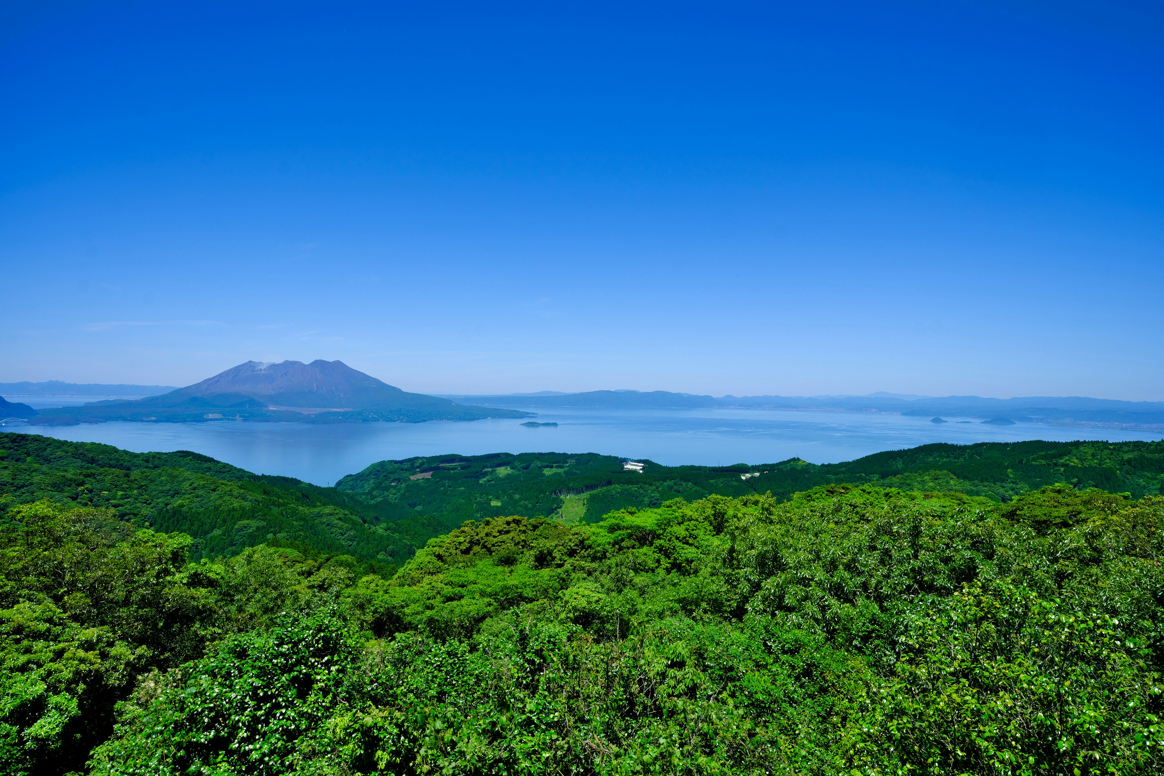 Pemandangan perbukitan hijau subur dengan gunung berapi di kejauhan di bawah langit biru cerah