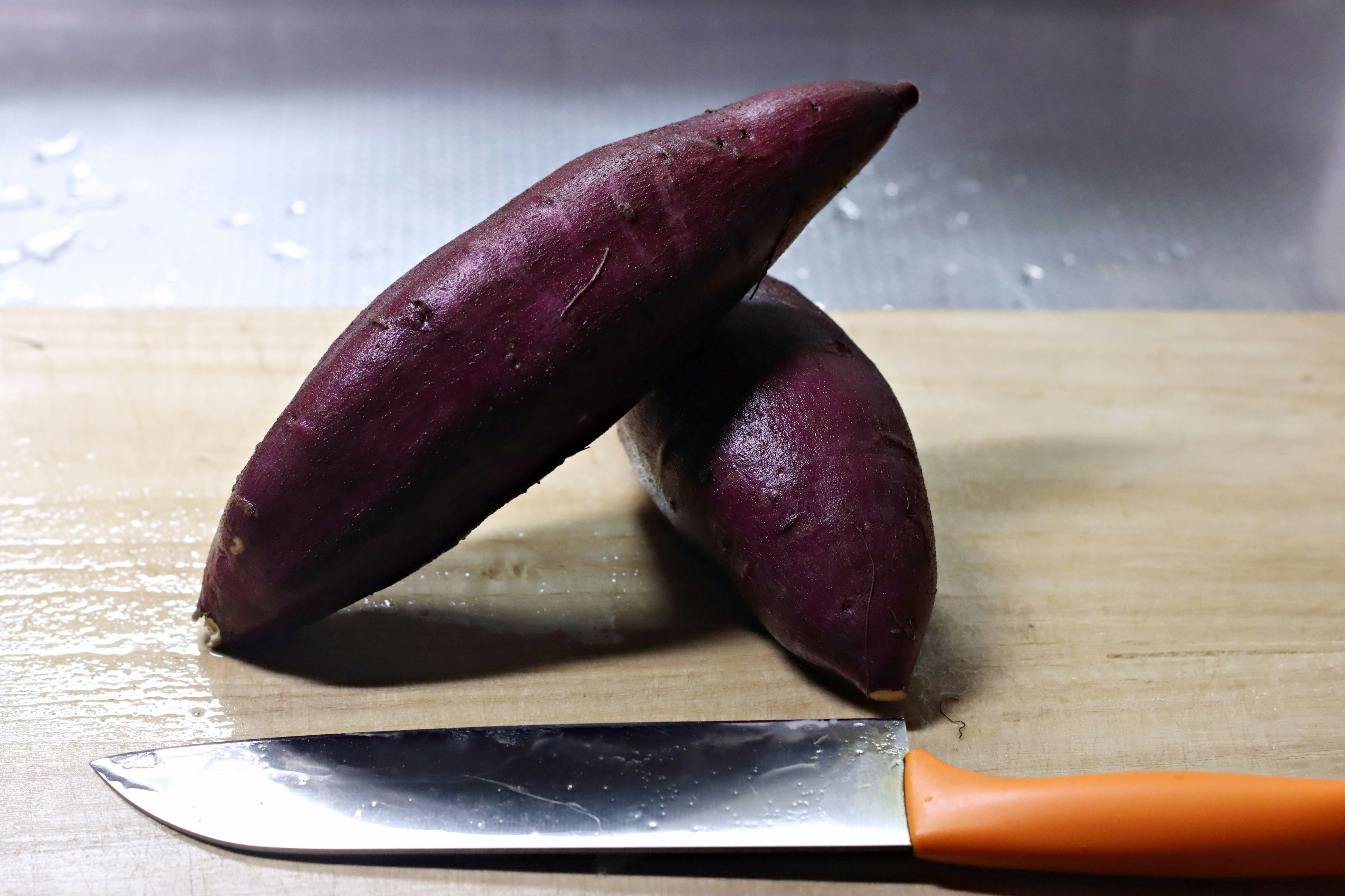 Two purple sweet potatoes on a wooden cutting board with an orange knife nearby