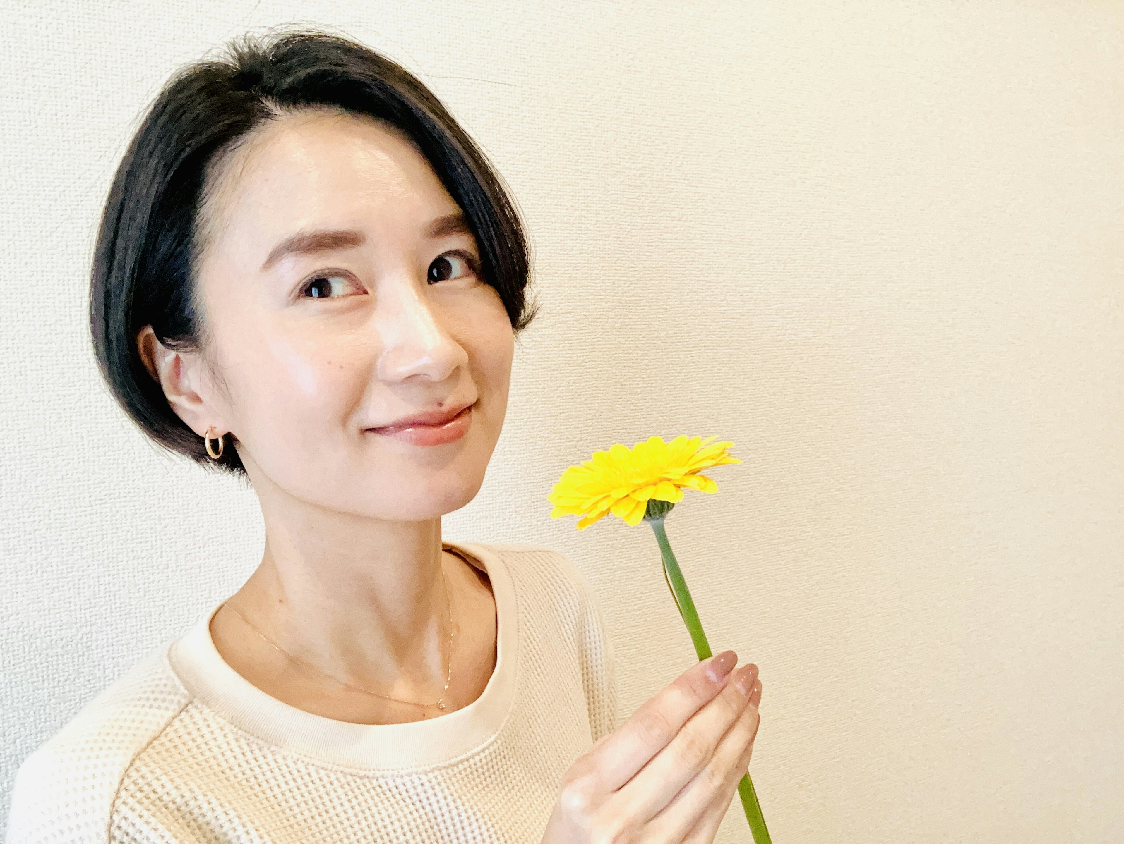 A woman smiling while holding a yellow flower against a simple background