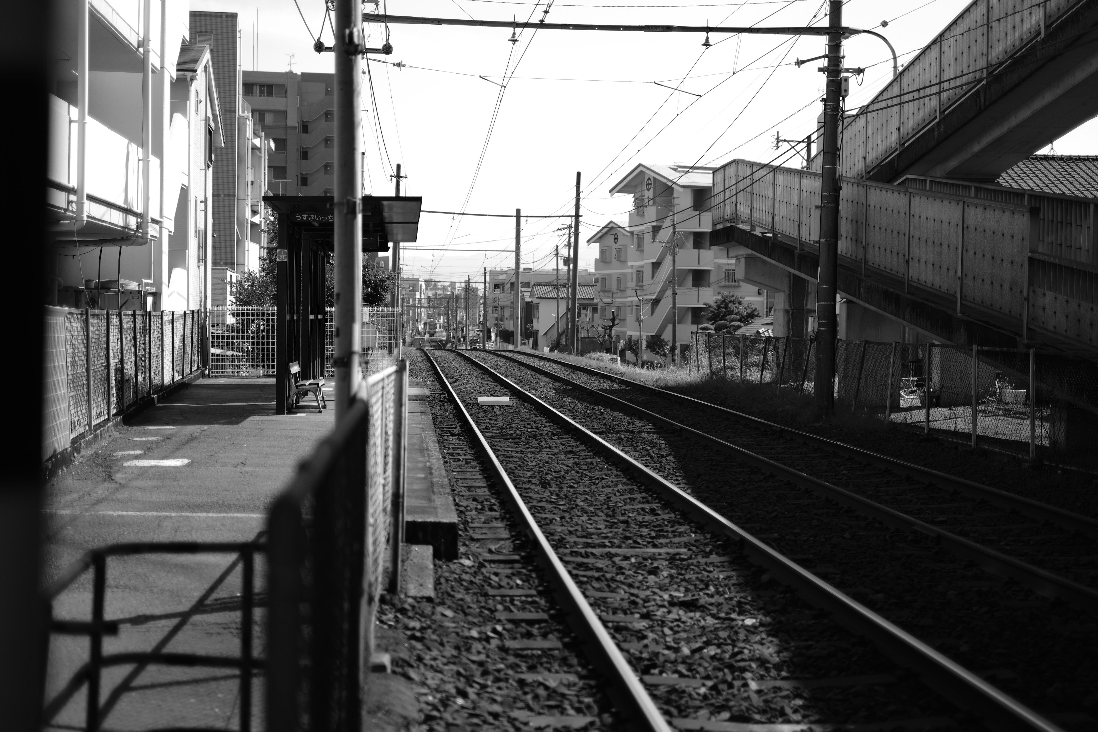 Scène ferroviaire en noir et blanc avec des voies ferrées et un quai de gare bâtiments à proximité et un viaduc