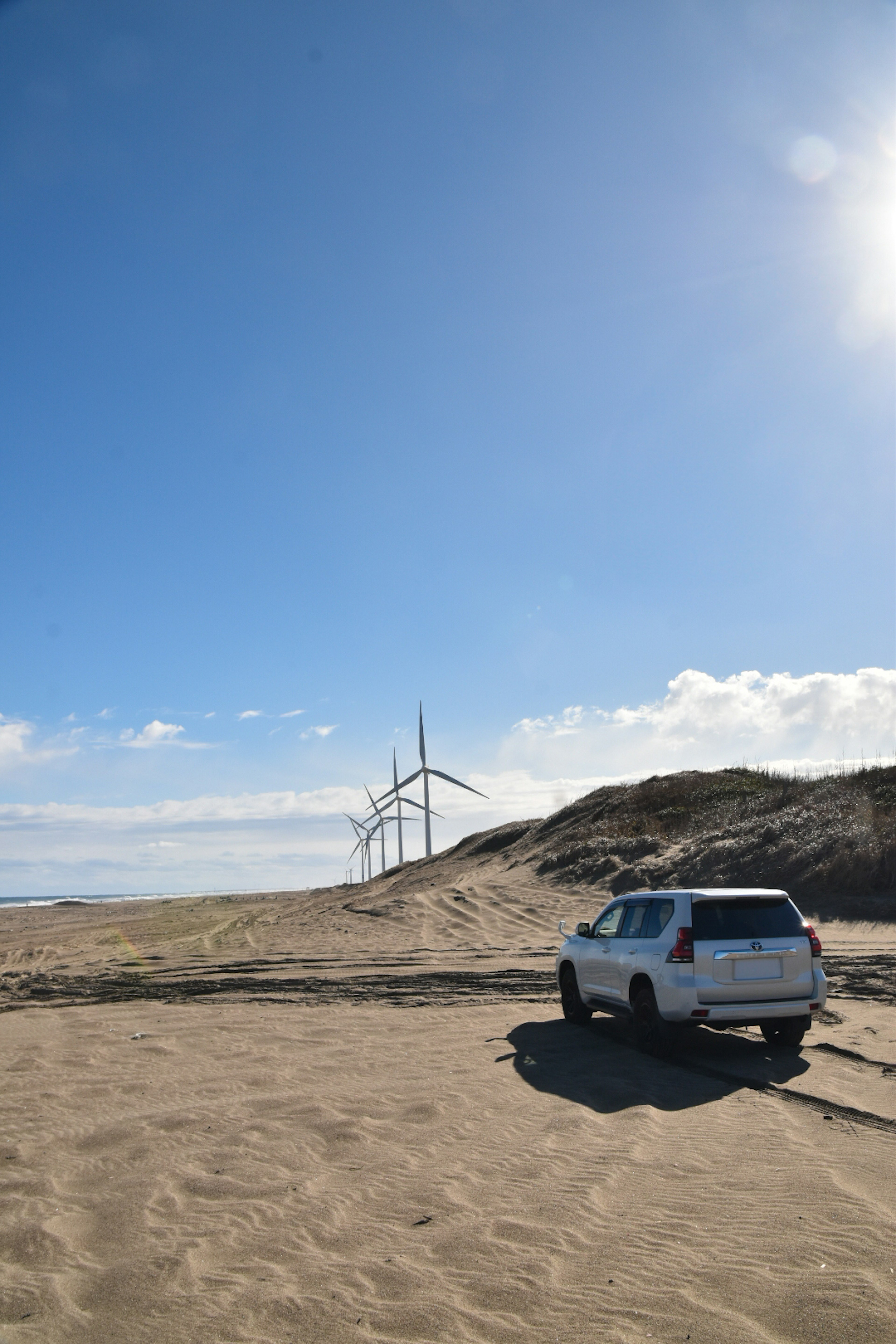 Un SUV blanco estacionado en una playa de arena con turbinas eólicas al fondo