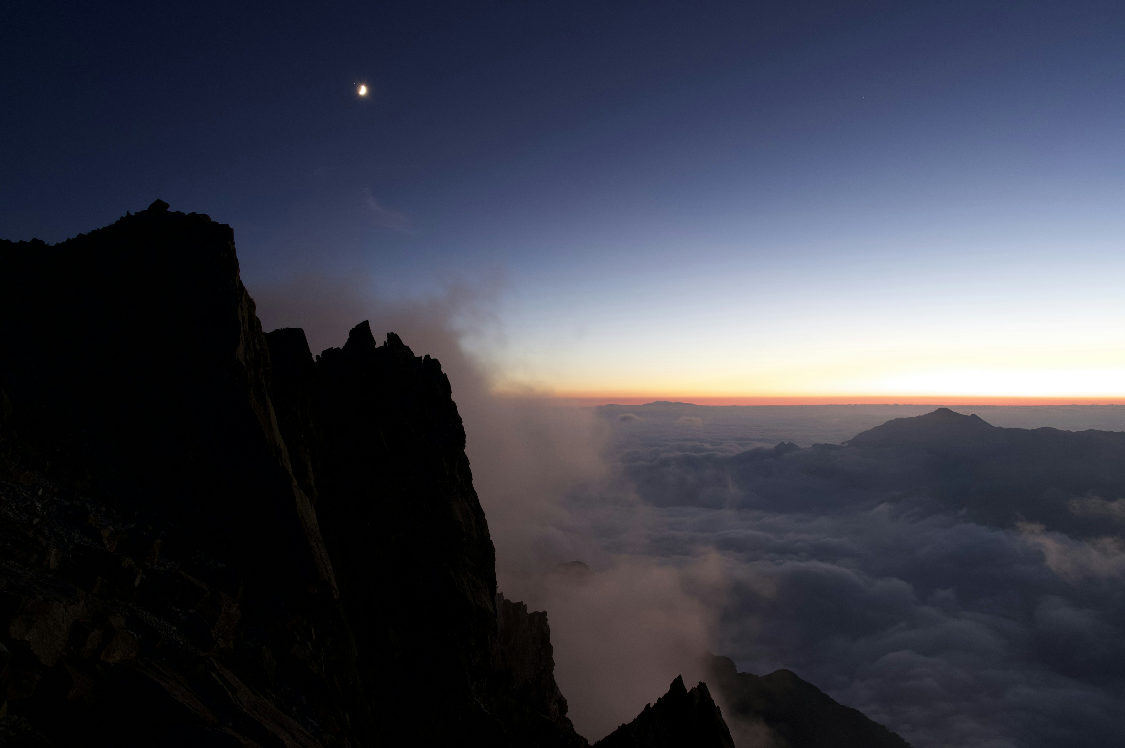 Silueta de montañas al anochecer con una luna sobre el mar de nubes