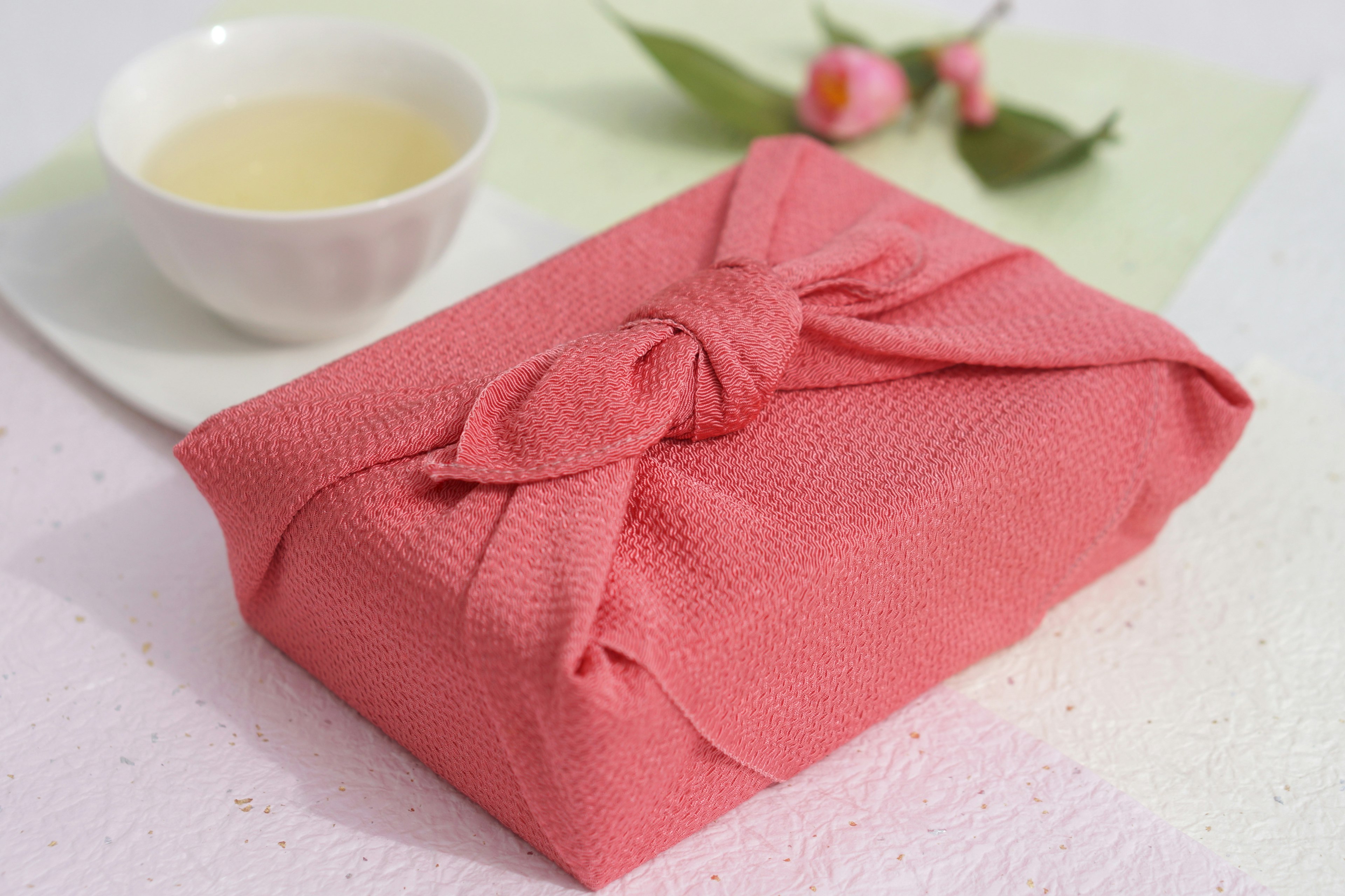 A pink fabric-wrapped gift beside a white tea bowl and green leaves