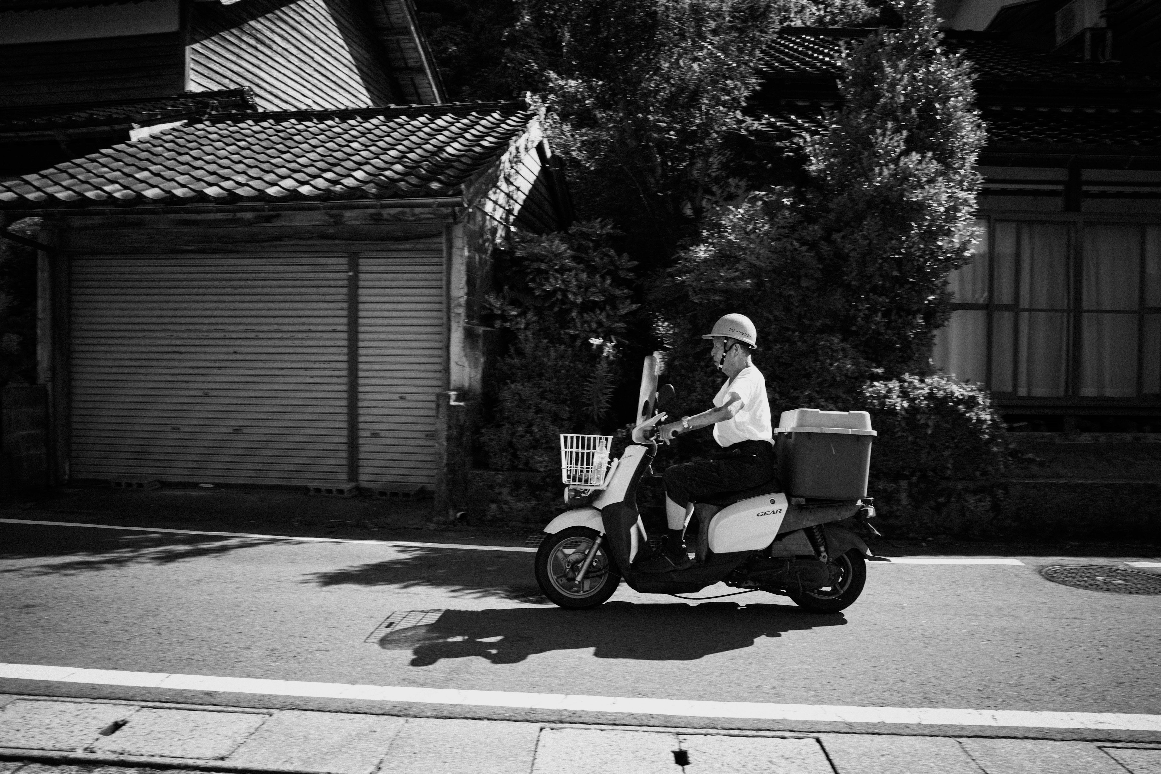 Un homme montant un scooter de livraison dans une rue en noir et blanc