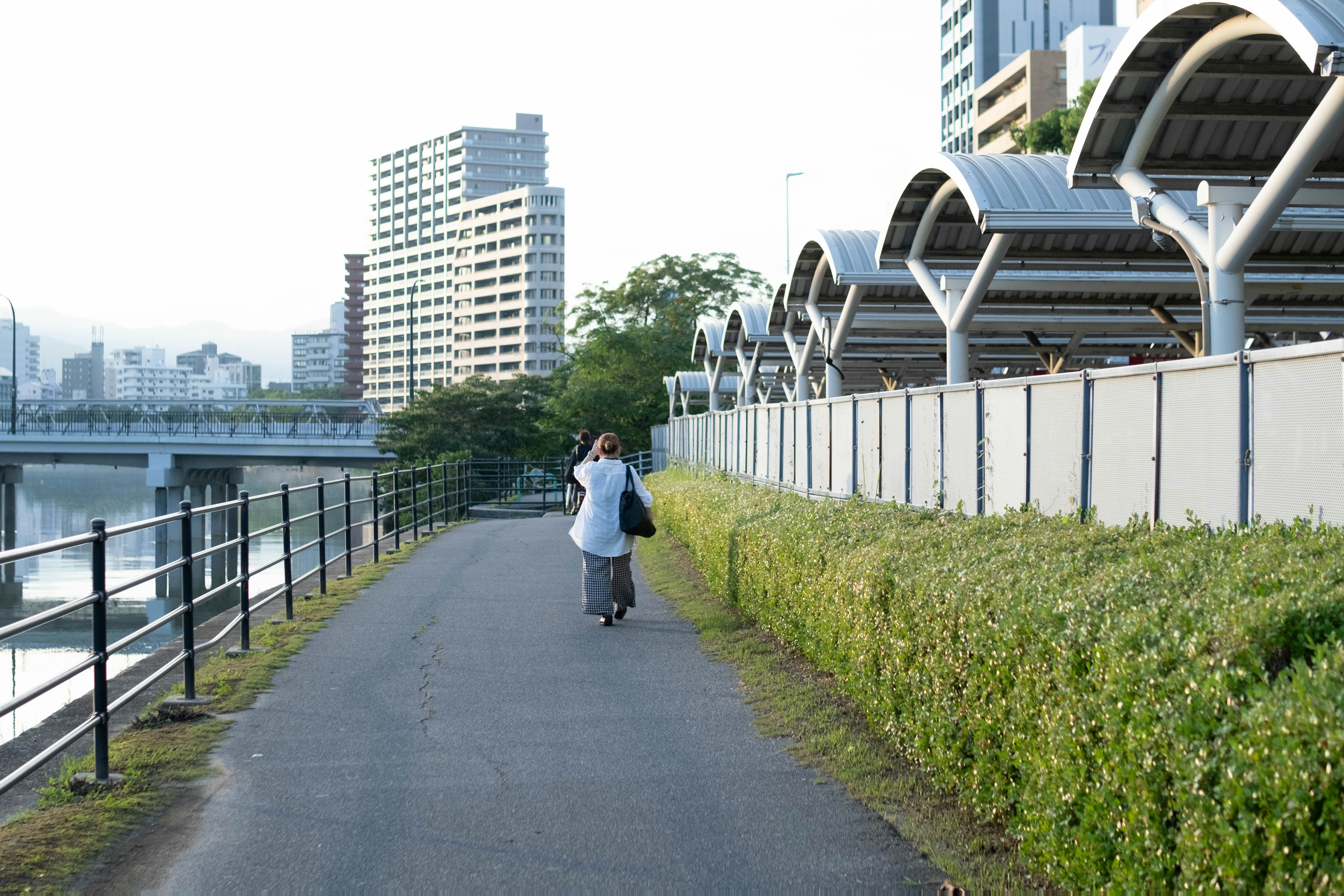 人が川沿いの道を歩いている風景 建物と緑のヘッジが見える