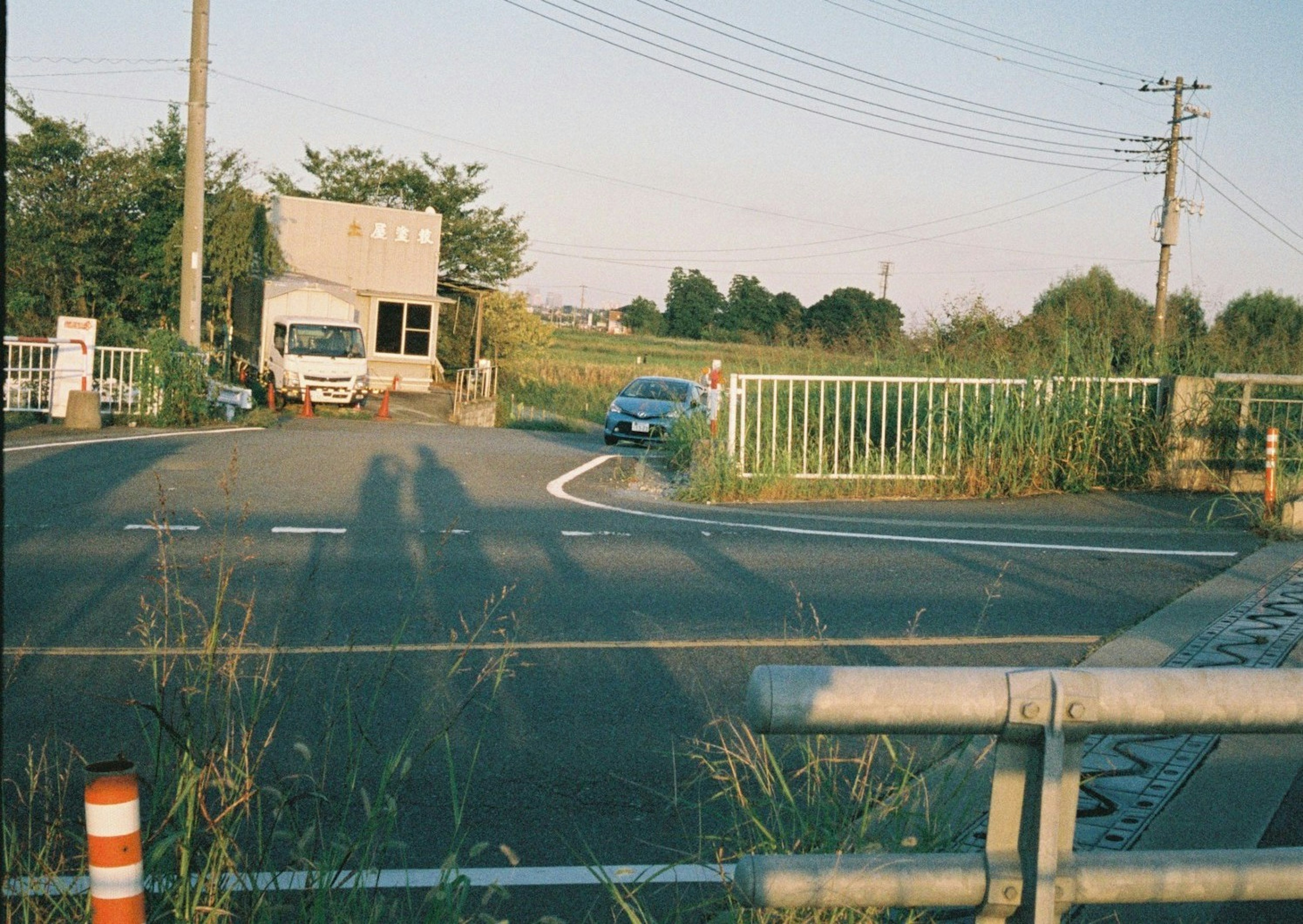 青い車が交差点に向かっている風景、背景にはトラックとフェンスがある