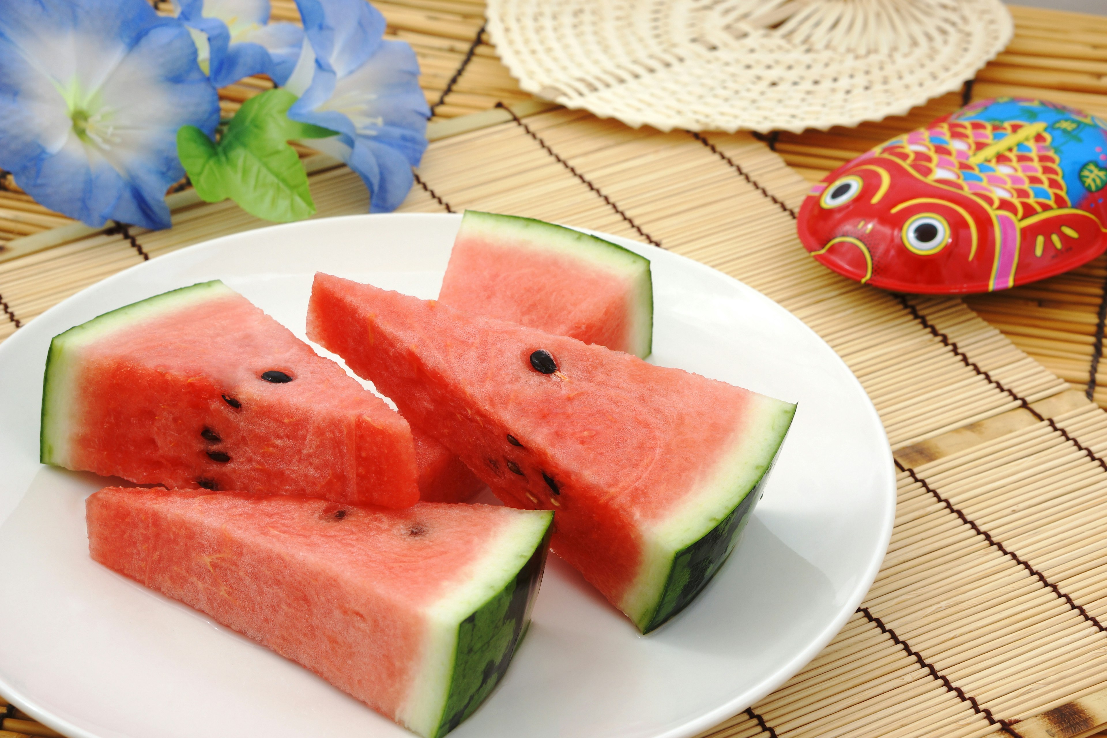 ชิ้นส watermelon วางอยู่บนจานสีขาวพร้อมดอกไม้สีน้ำเงินและการตกแต่งปลาสีสันสดใสในพื้นหลัง